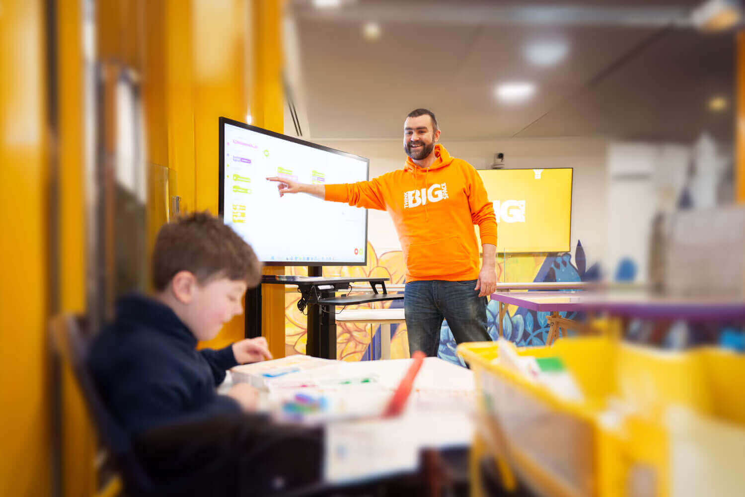 A teacher in an orange shirt engages students in a colorful classroom with interactive learning activities.