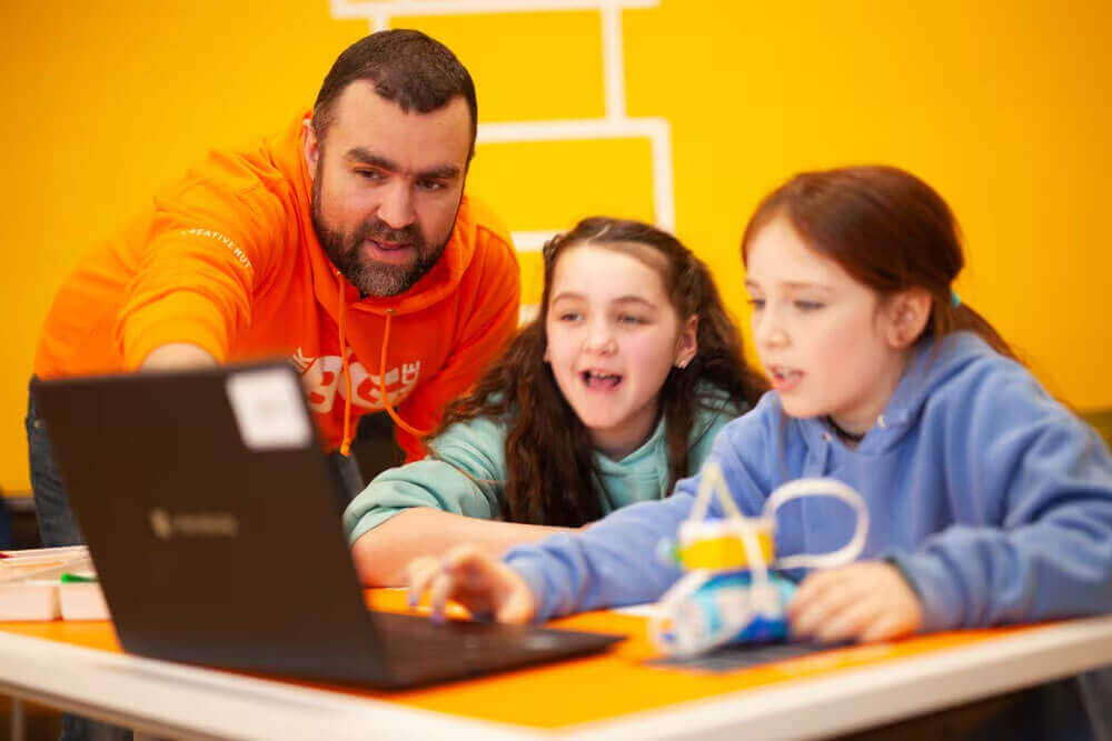 Teacher guiding two engaged students at a table with a laptop, fostering a learning environment.