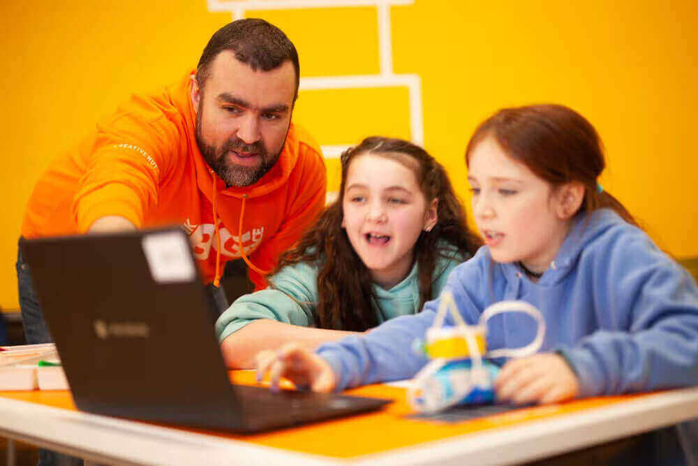 A mentor helps two children with a laptop in a bright, engaging learning environment, showcasing teamwork and education.