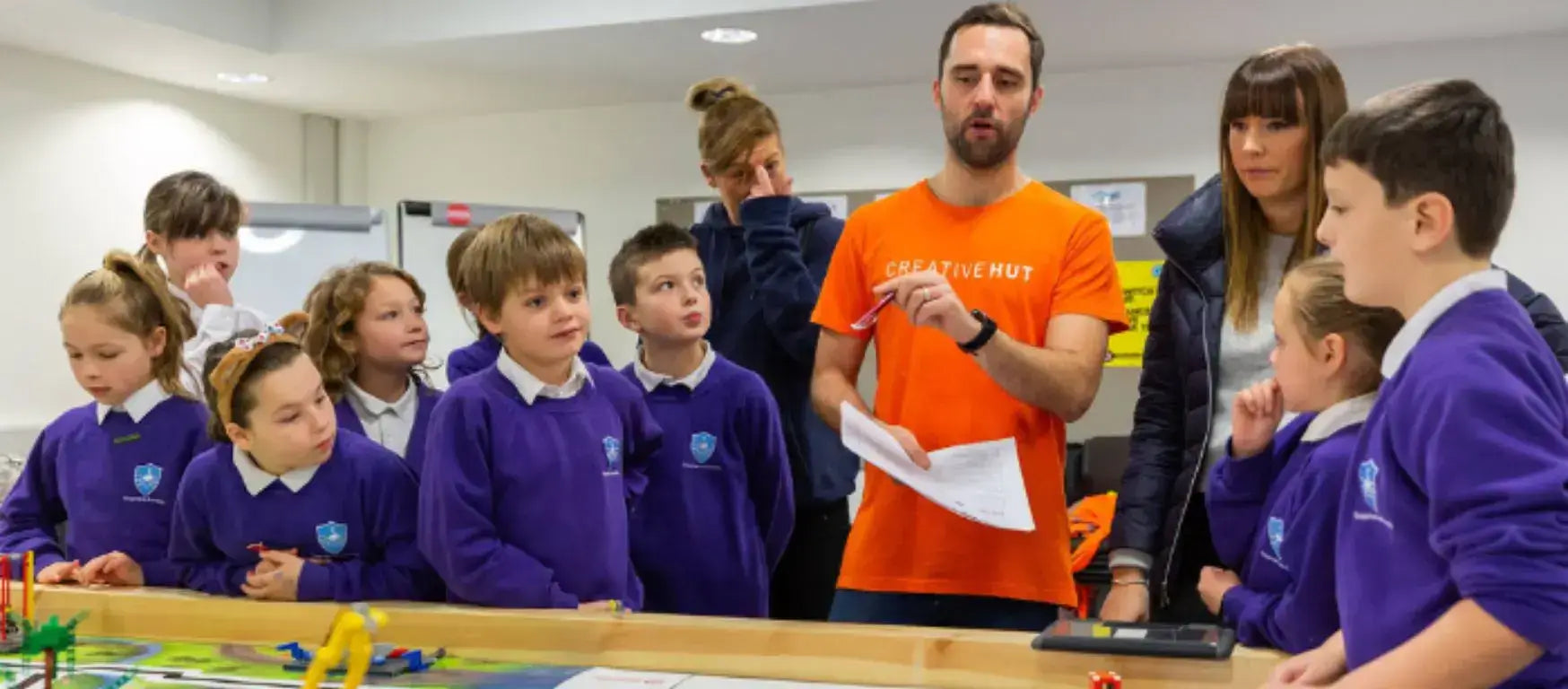 A CreativeHUT instructor engages with children in purple uniforms during a STEM lesson involving robotics.