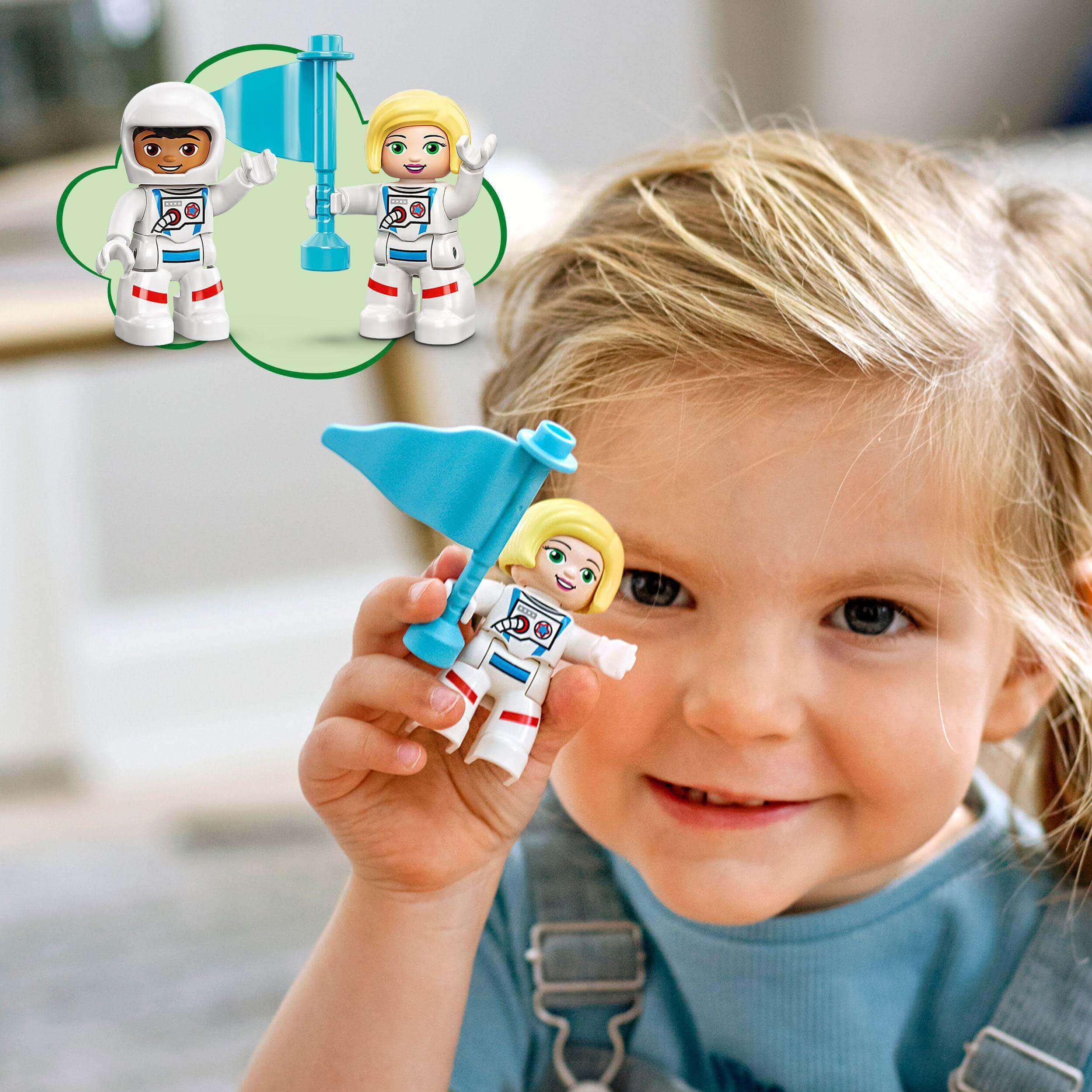 A toddler smiling while holding a LEGO® DUPLO astronaut figure and blue flag, showcasing imaginative play.