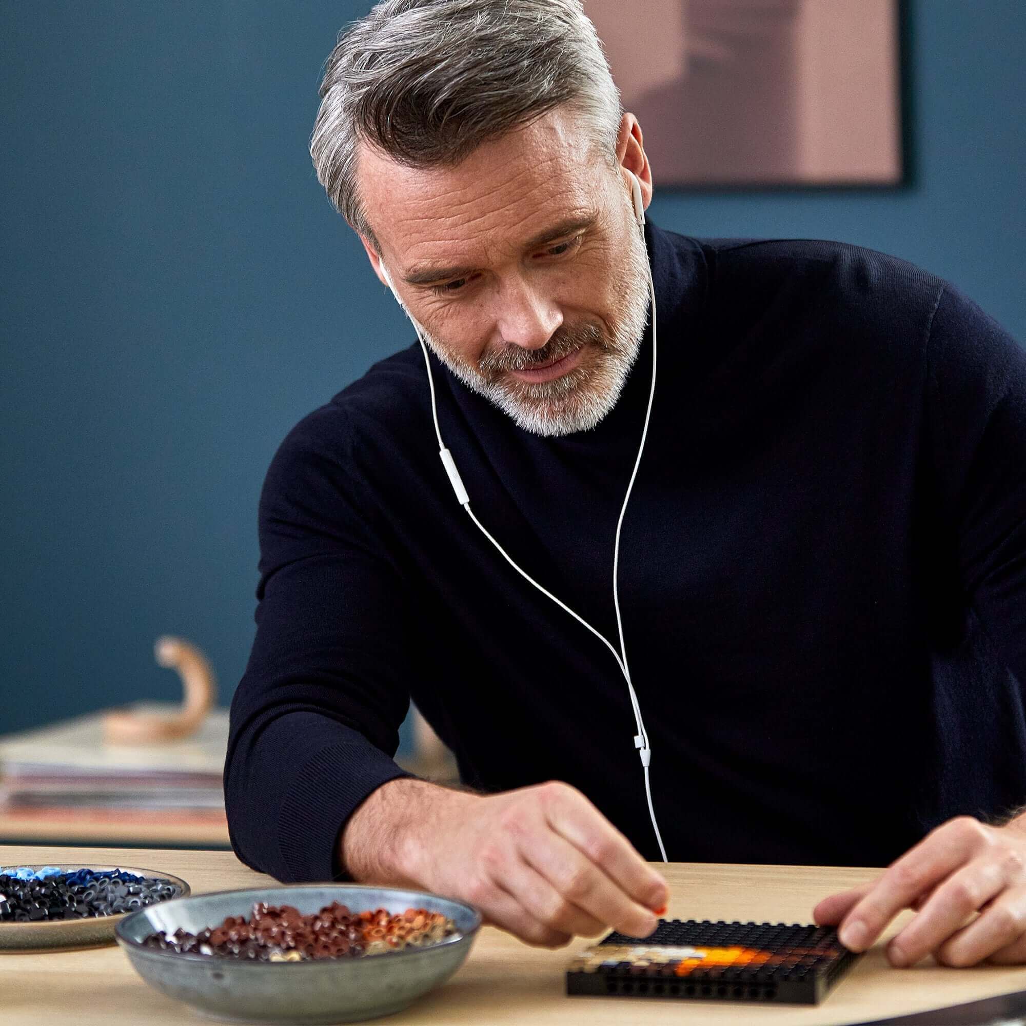 Adult man building LEGO® Art The Beatles set, enjoying immersive soundtrack, with colorful brick pieces and bowls on table.