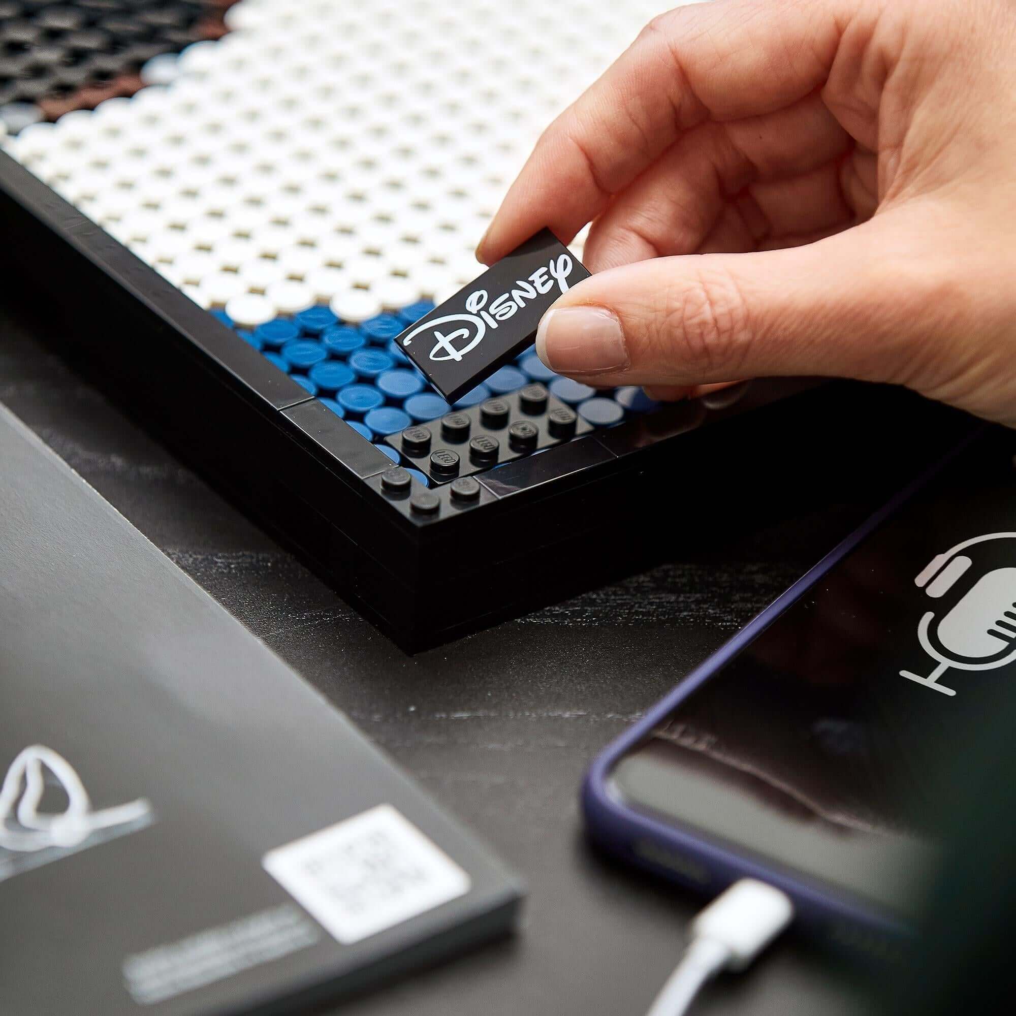 Hand placing a LEGO® tile featuring Disney logo on Mickey Mouse Poster Set 31202 during assembly.