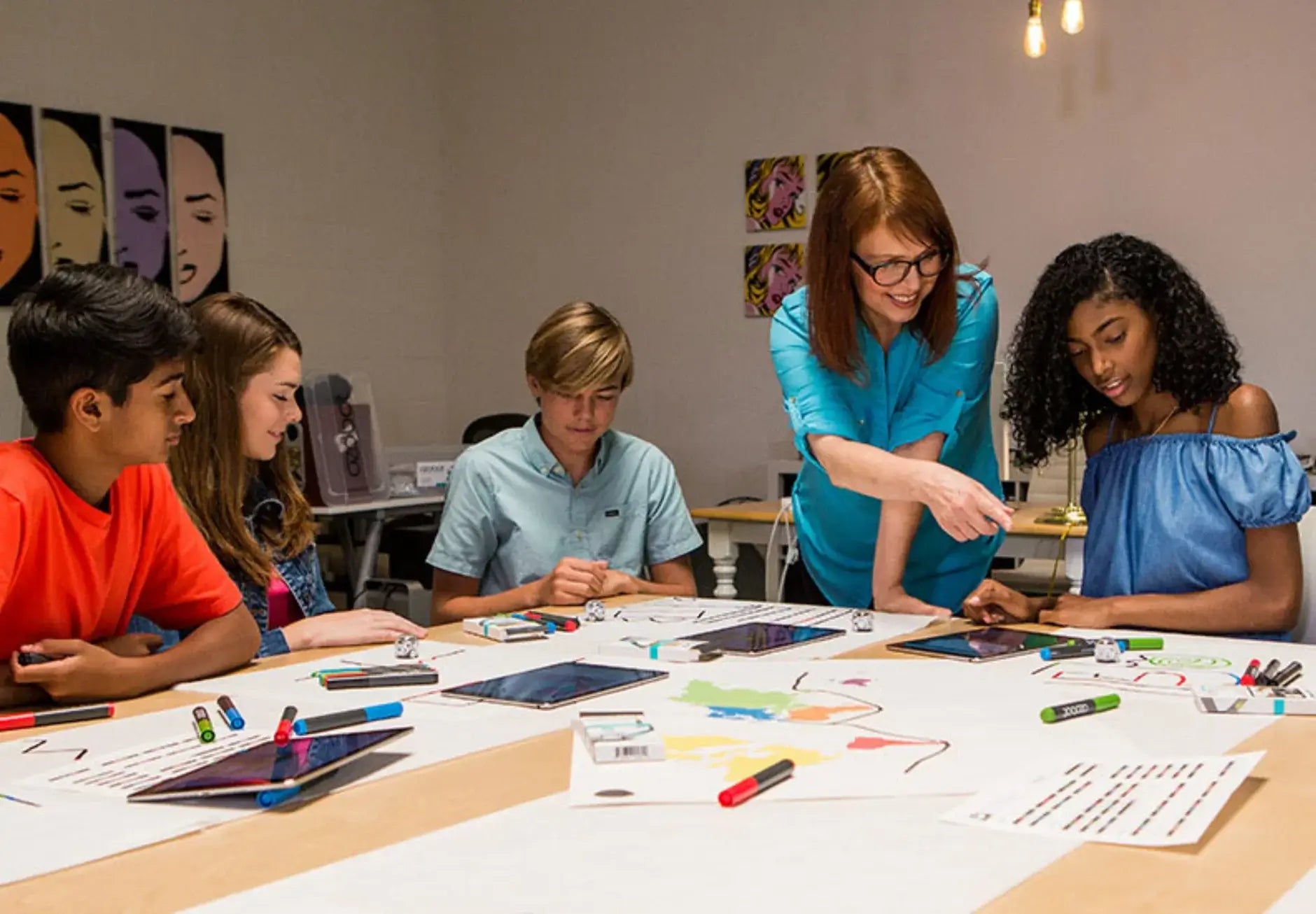 A teacher assists students with art projects using tablets and markers in a creative classroom setting.