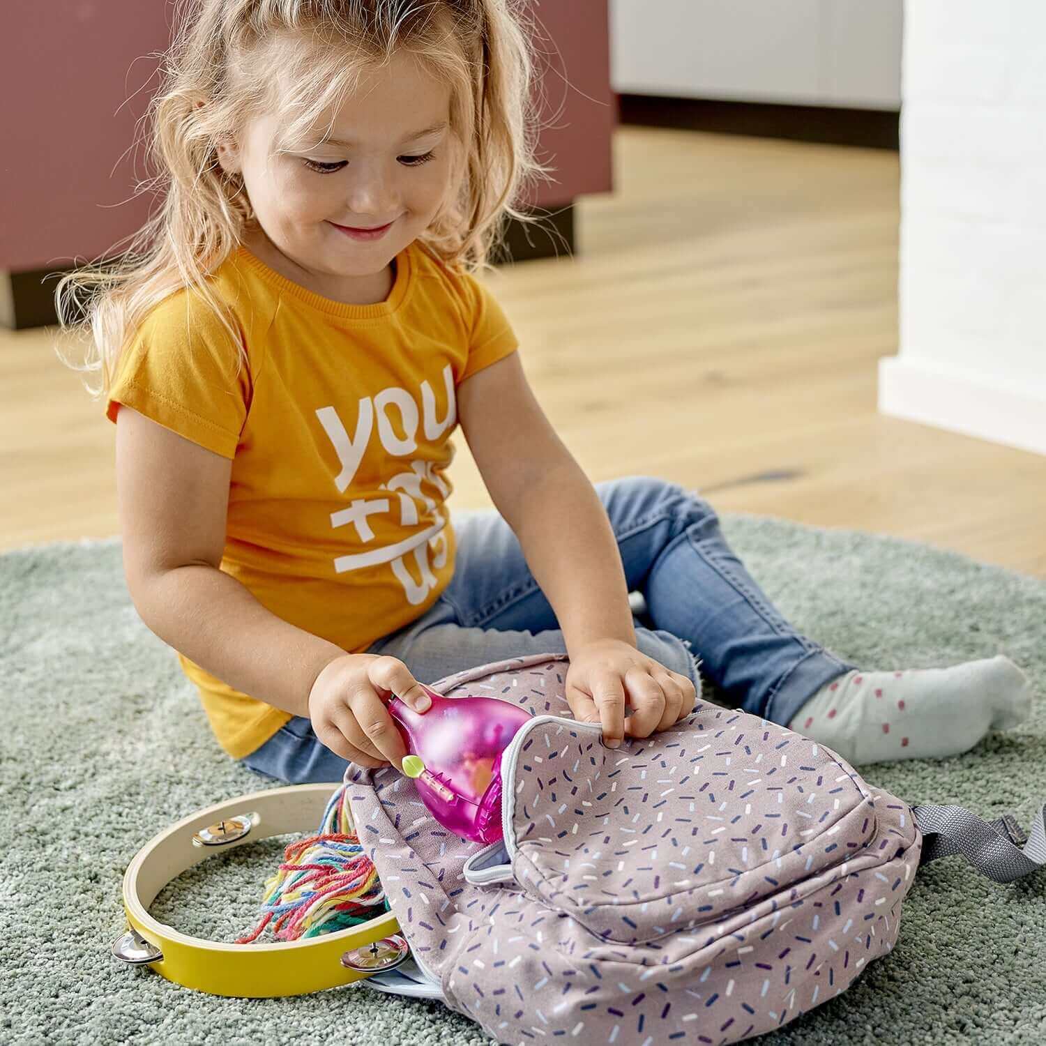 Young girl unpacking LEGO® Trolls Poppy's Pod Storage set from colorful backpack on a cozy rug.
