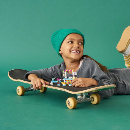 Child smiling on a skateboard, playing with a colorful LEGO® Friends Ice Cream Cart set against a green background.