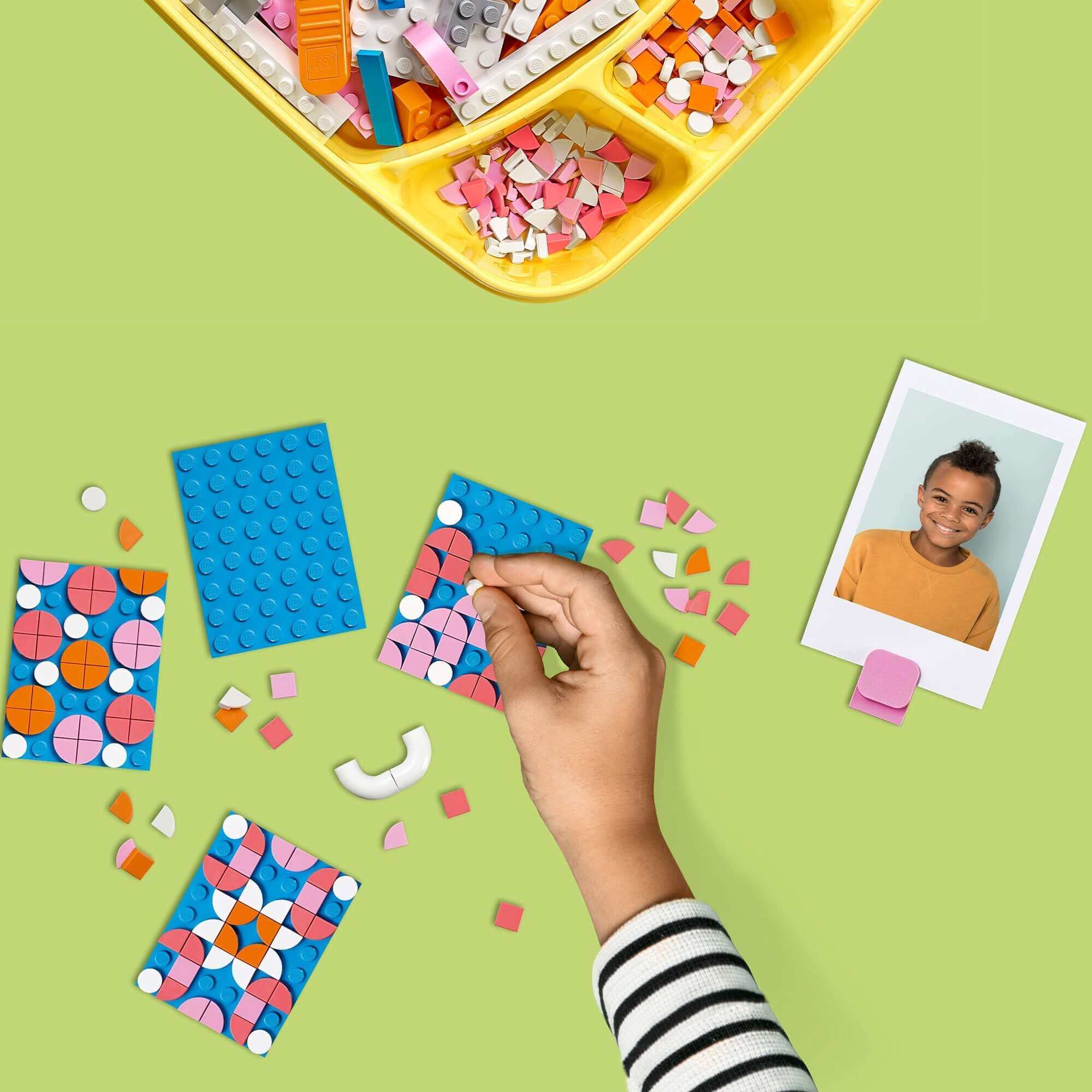 Person decorating LEGO® DOTS with colorful tiles, featuring a photo holder and craft elements on a vibrant desk.