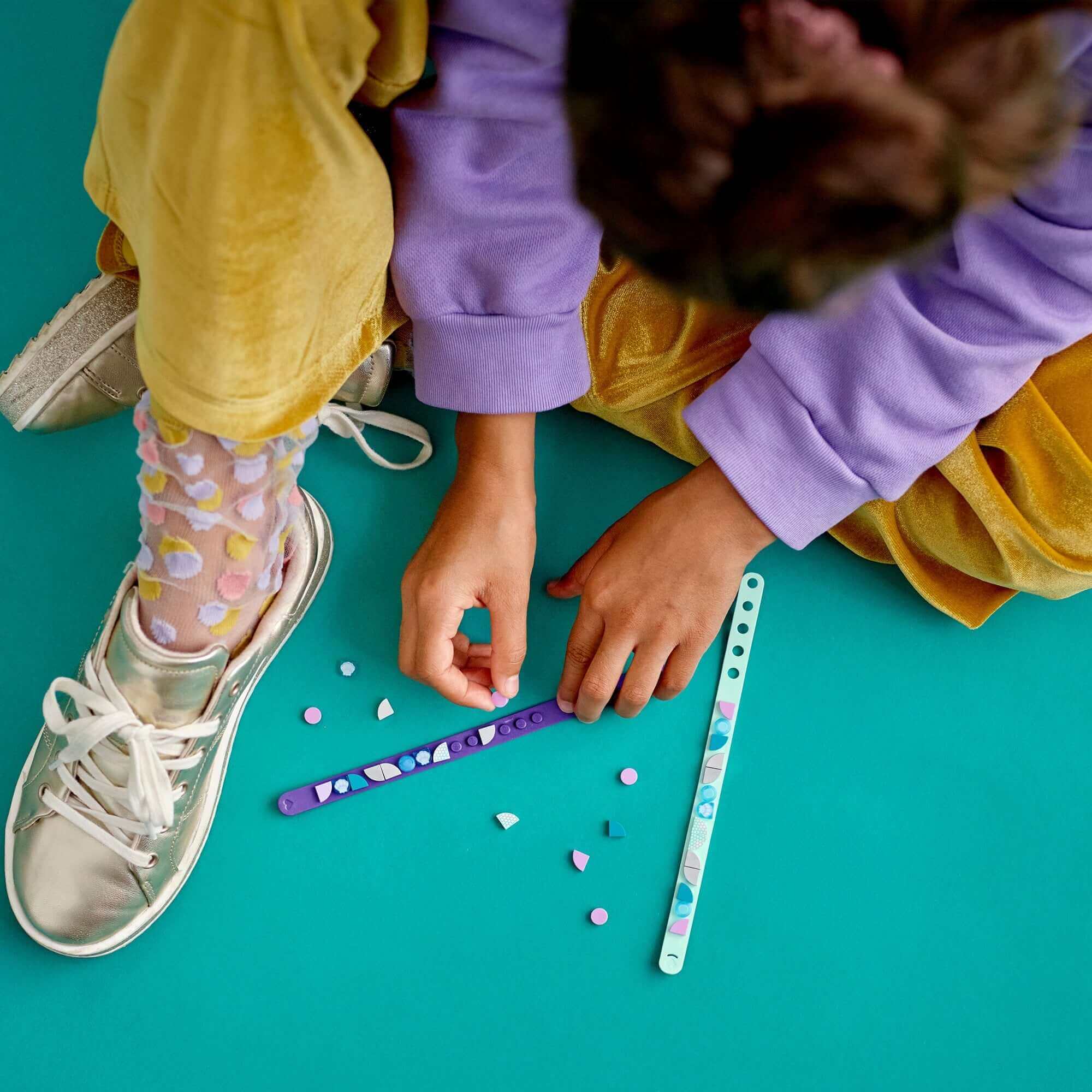 Child crafting with LEGO® DOTS Mermaid Vibes Bracelets, colorful tiles on teal background.