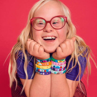 Child wearing colorful LEGO® DOTS Mermaid Vibes bracelets, smiling with joy against a vibrant pink background.