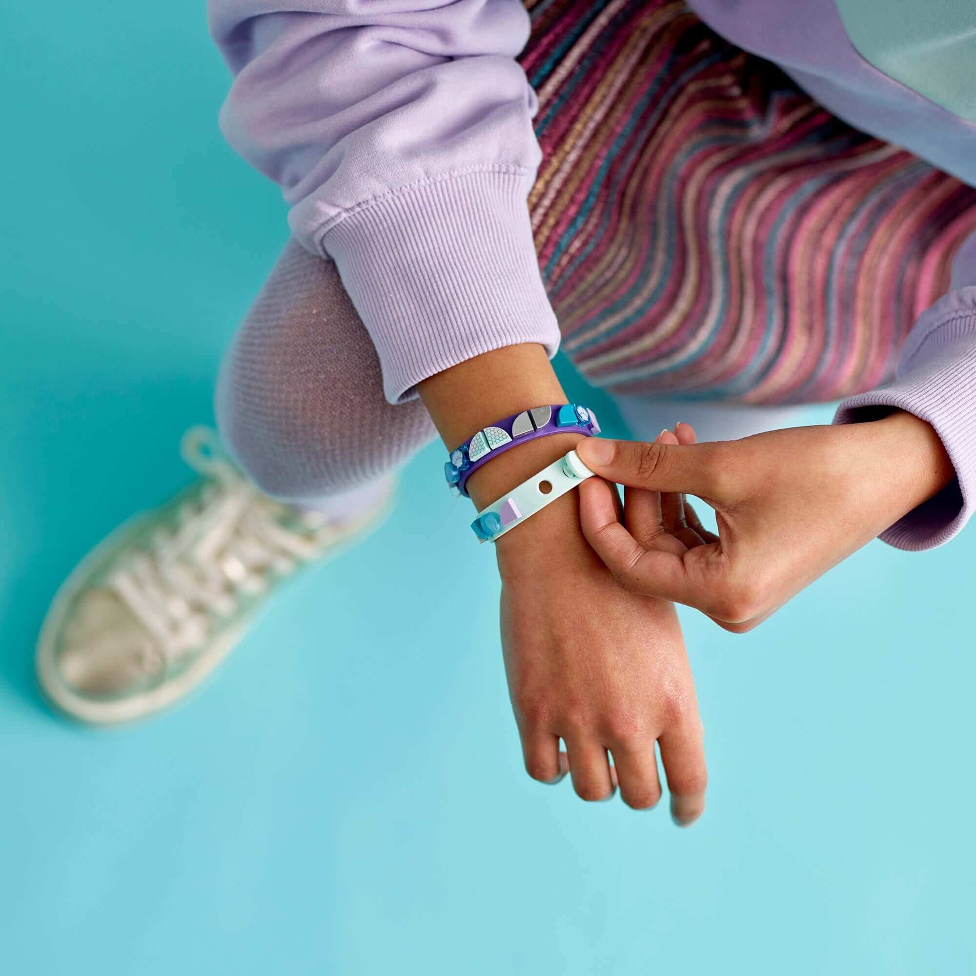 Child wearing LEGO® DOTS Mermaid Vibes Bracelets with colorful tiles on a blue background, showcasing creativity.