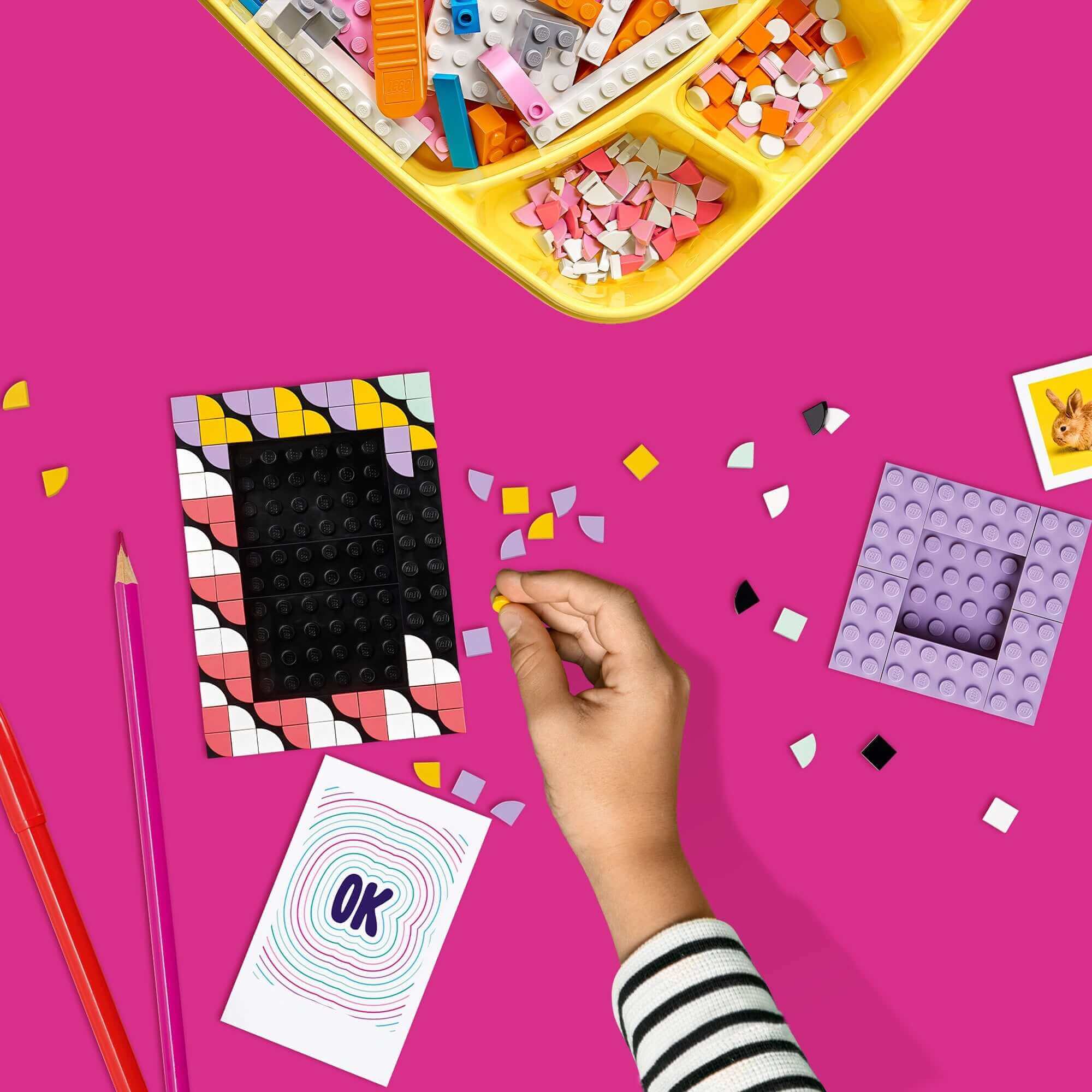 Child's hand creating designs with LEGO® DOTS picture frames and colorful tiles on a bright pink background.