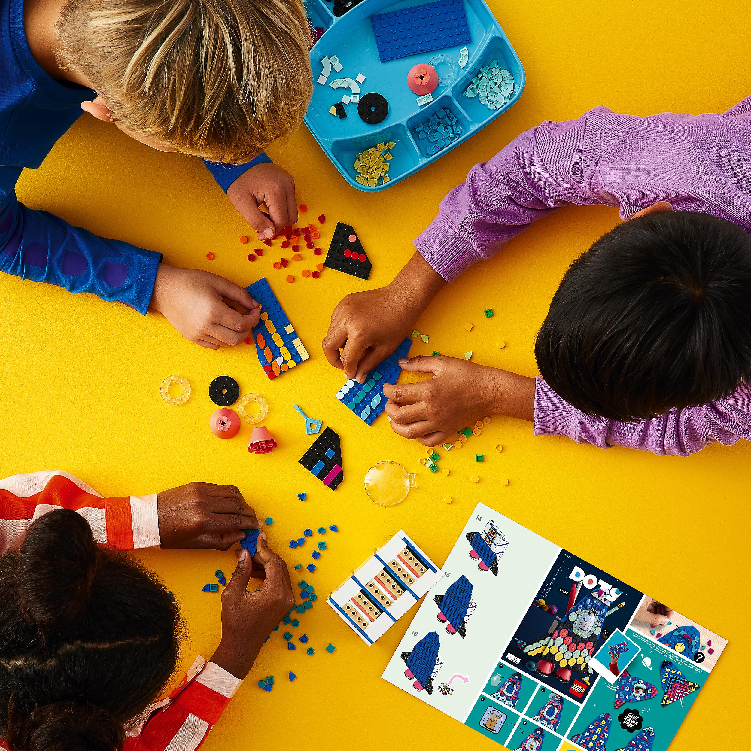 Kids creating colorful designs with LEGO® DOTS Lots of DOTS Tiles Craft Set on a yellow surface, showcasing creativity.