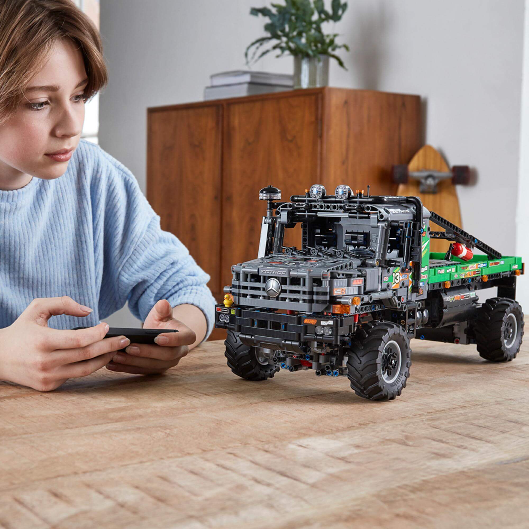 Child controlling LEGO® Technic 4x4 Mercedes-Benz Zetros Truck with smartphone on wooden table.