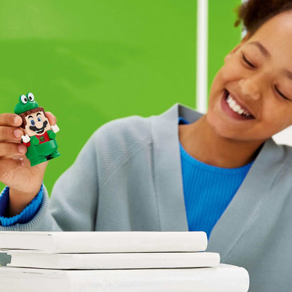 Child holding LEGO® Super Mario Frog Mario figure, smiling with books in the foreground, vibrant green background.