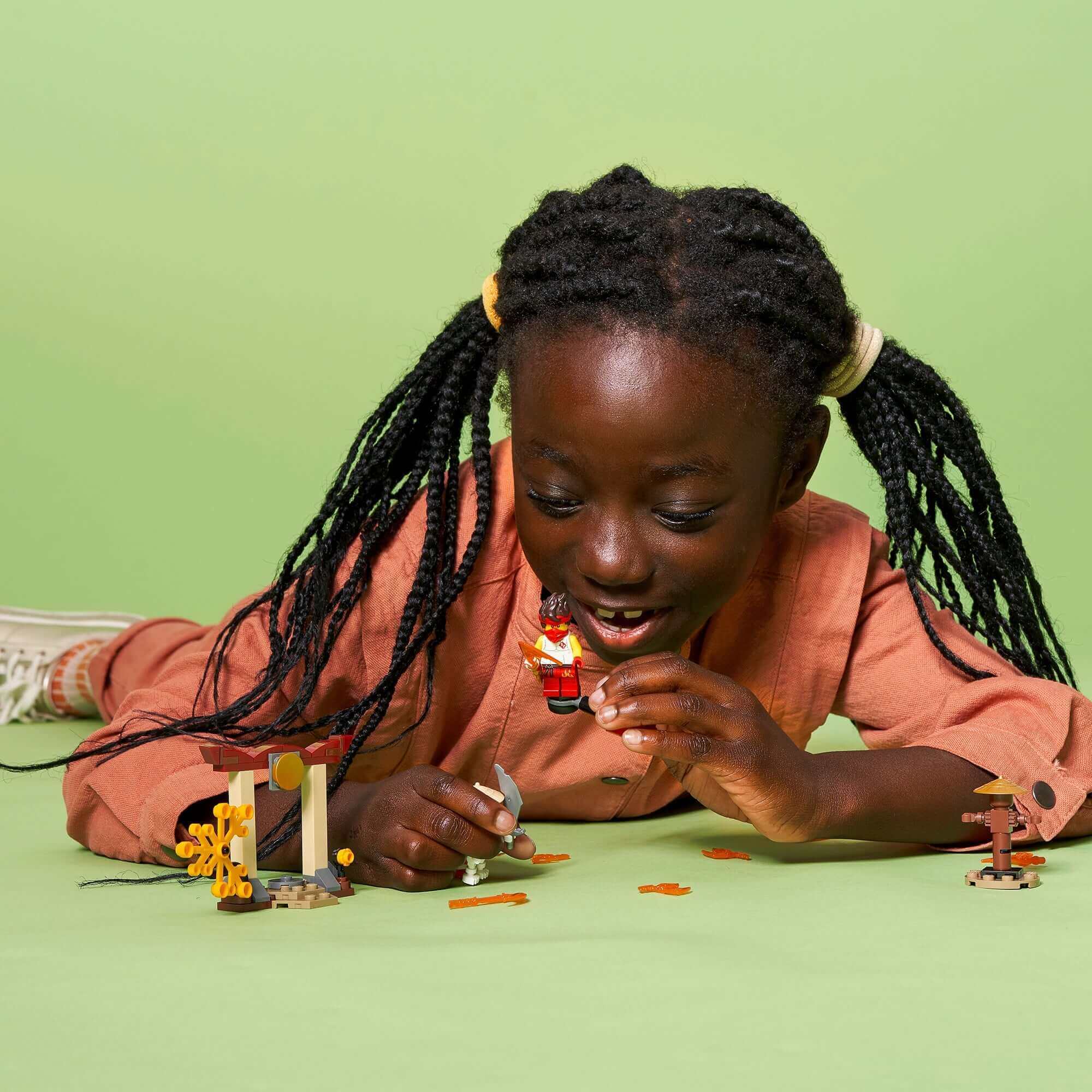 Child playing with LEGO® NINJAGO set, enjoying Kai vs. Skulkin battle on a green background.