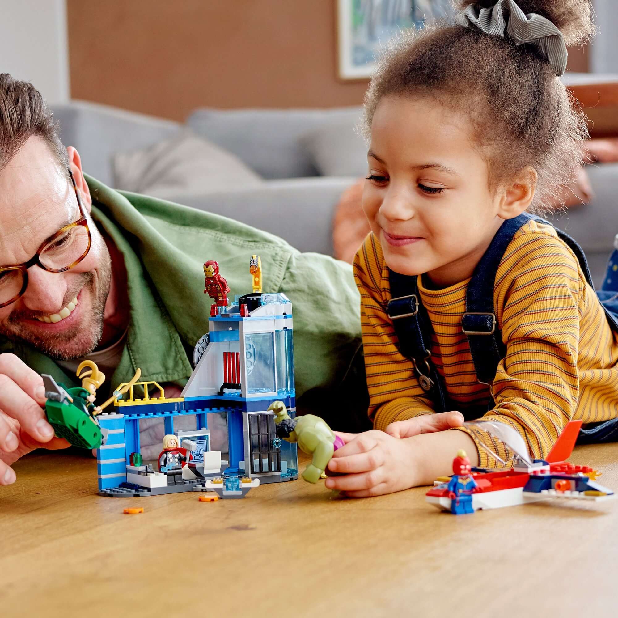 A father and daughter enjoy playing with the LEGO® Marvel Avengers Wrath of Loki set 76152 on a wooden floor.