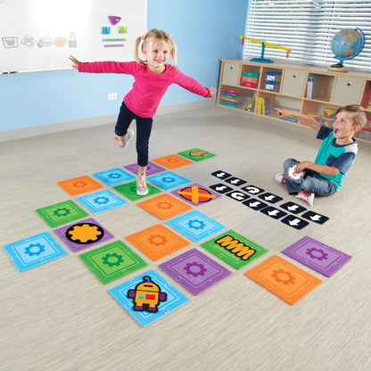 Two children playing with a life-size coding maze. One child balances on a foam mat while the other points and holds a coding card, demonstrating active, screen-free programming.