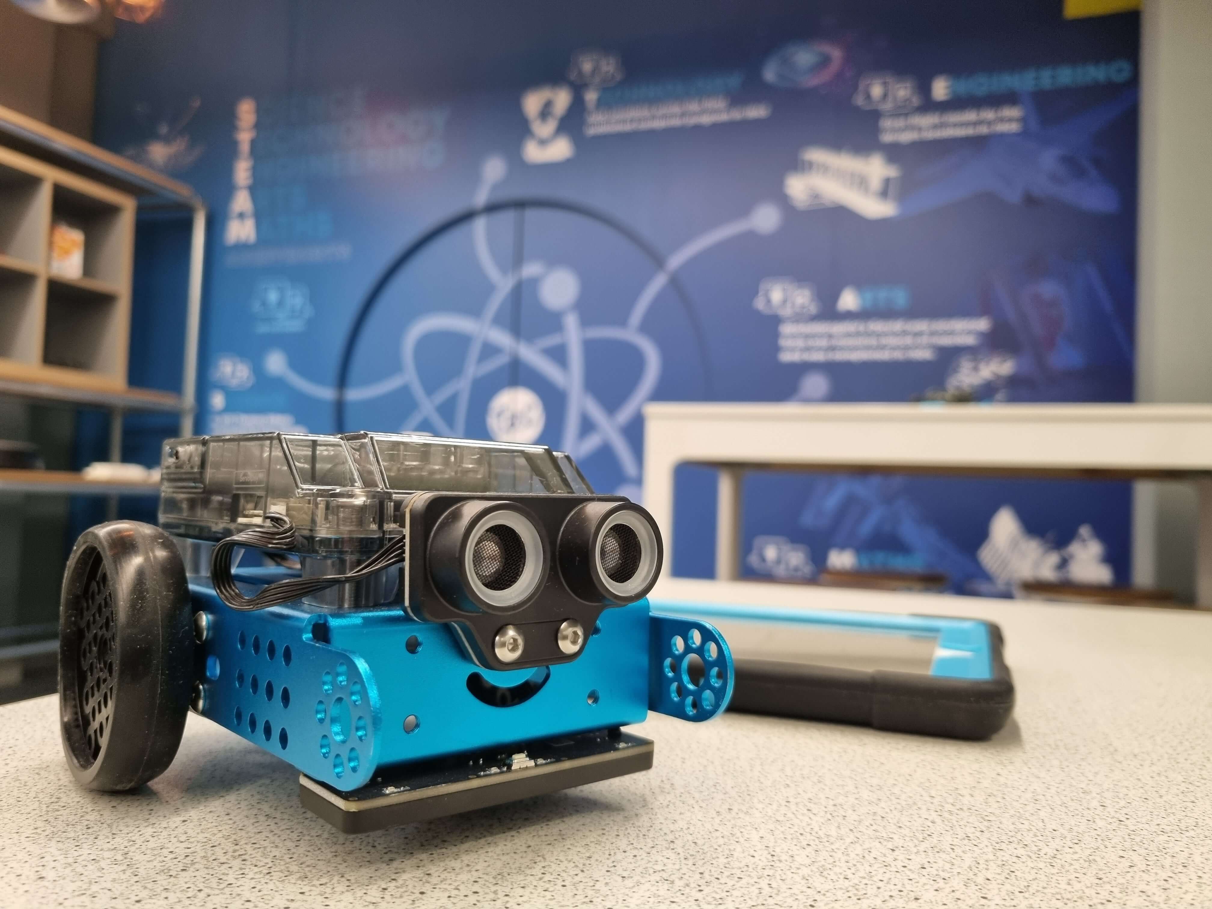 A playful blue robot with wheels and smiling face, displayed in a tech-themed educational environment.