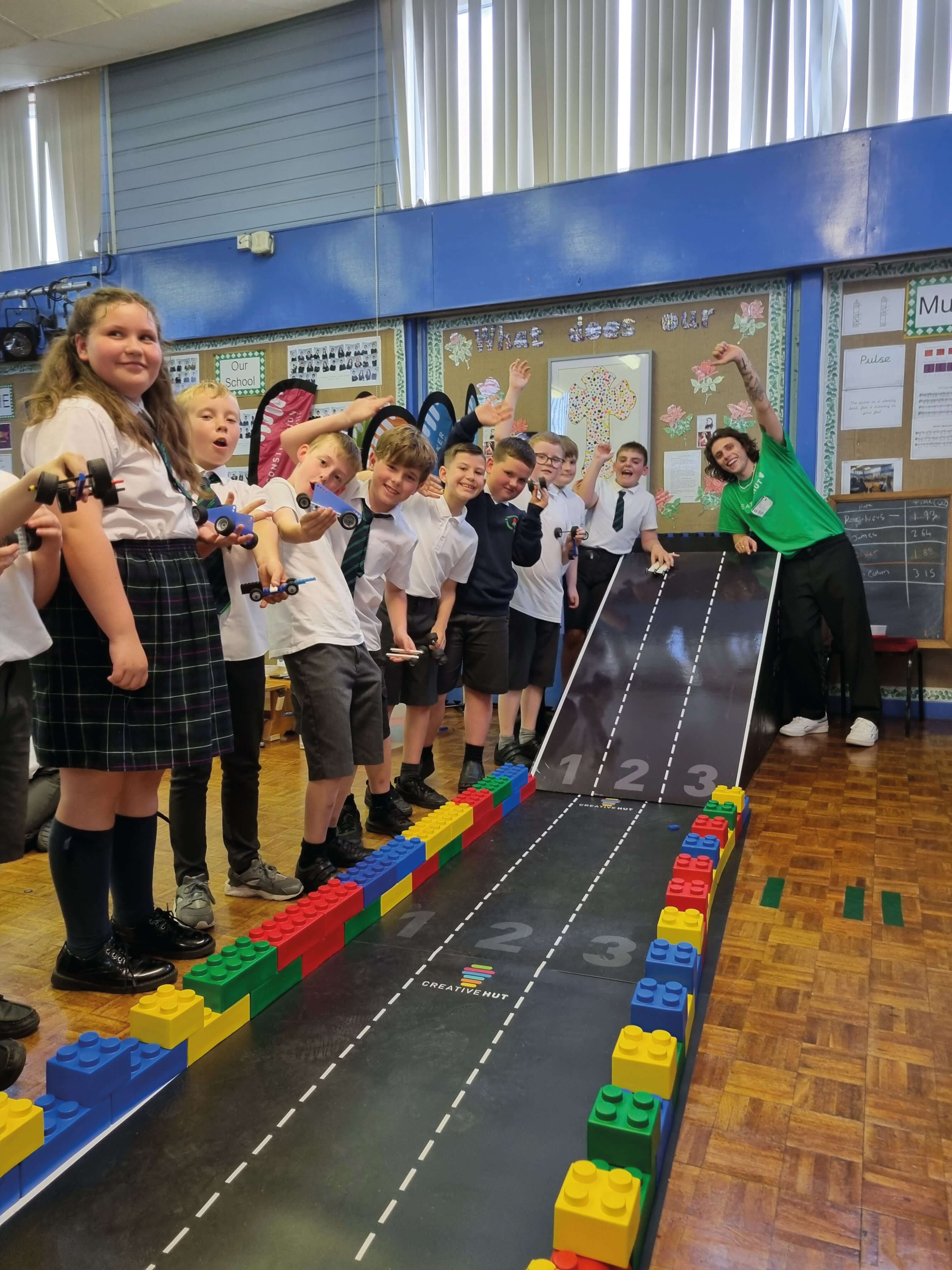 Students playing with toy cars on a colorful LEGO racetrack in a classroom, with a smiling teacher guiding them.