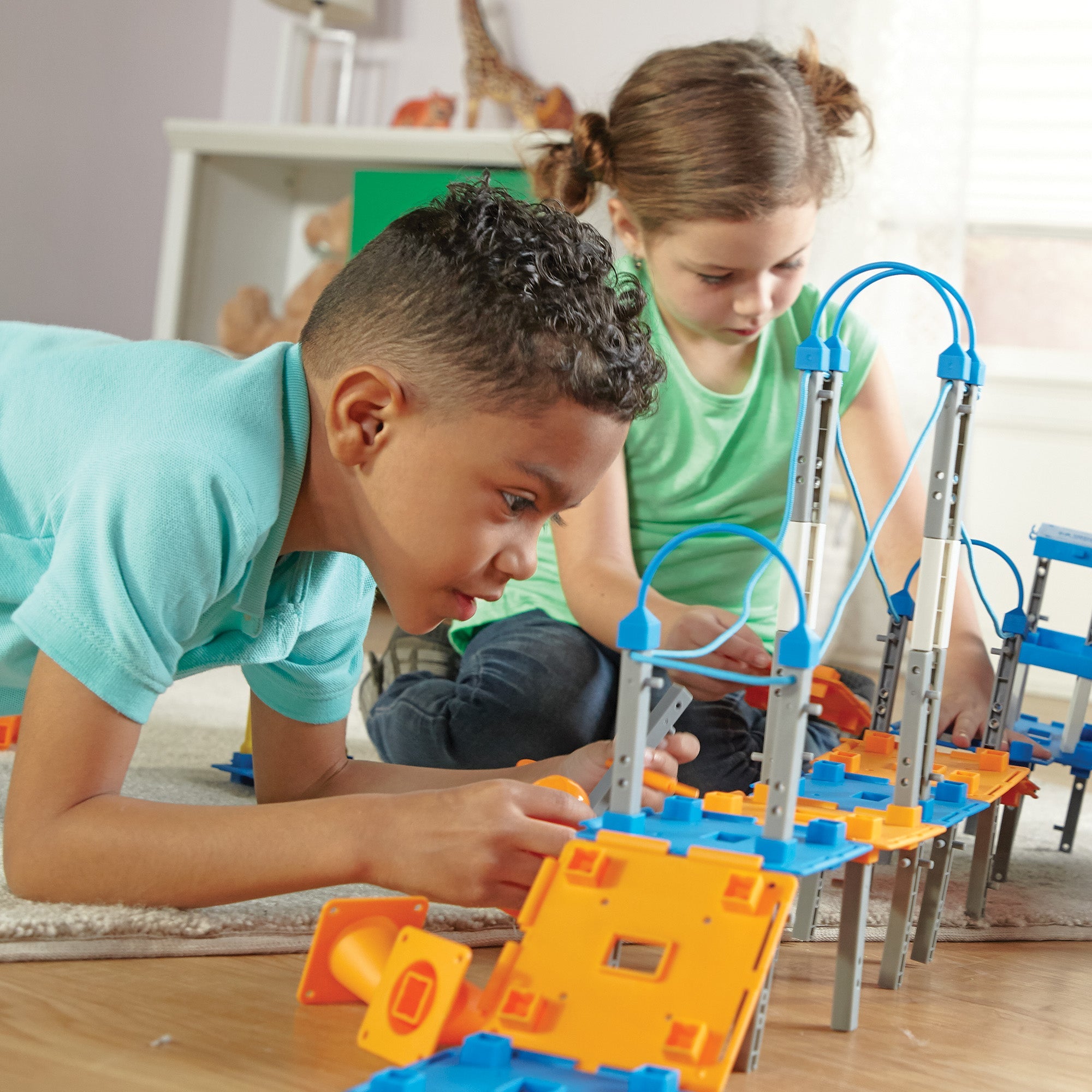 Two children collaborating on a floor to build a complex city structure. They are using blue and orange platforms and grey beams from a 100-piece STEM engineering set.