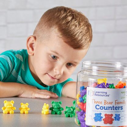 A young boy leans over a table, intently focused on sorting and lining up colourful Three Bear Family counters. In front of him, he has arranged several yellow and green bears by size. To the right, a large, clear storage jar labelled "Learning Resources Three Bear Family Counters" is filled with a vibrant mix of 96 bears in six different colours. The scene demonstrates hands-on learning, size-grading, and fine motor skill development in a home or classroom setting.