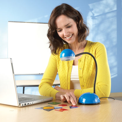 A teacher in a yellow cardigan sits at a desk, smiling as she uses the Luna™ 2.0 Interactive Projection Camera to demonstrate a maths activity. The blue, flexible gooseneck camera is positioned over several colourful, translucent geometric shapes on the desk. A laptop is connected to the camera via USB, showing how the device shares live "manipulatives in action" with the class. The background features a classroom setting with a whiteboard, illustrating the camera's use as a versatile multimedia teaching.