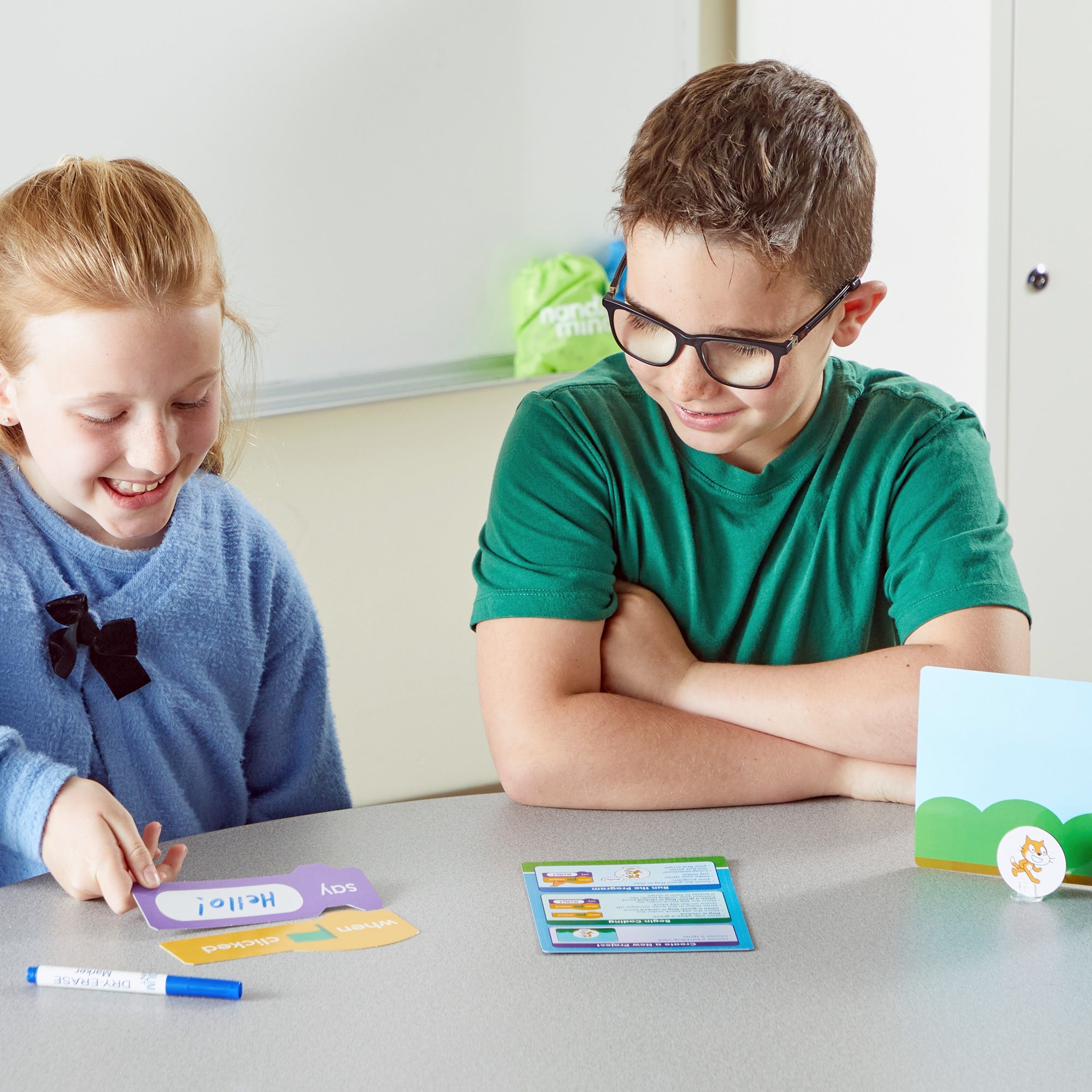 Two students smiling while using the Scratch Block-Based Coding Activity Set. They are arranging colourful, write-and-wipe coding tiles on a table to build a program sequence near character sprites and backdrops.