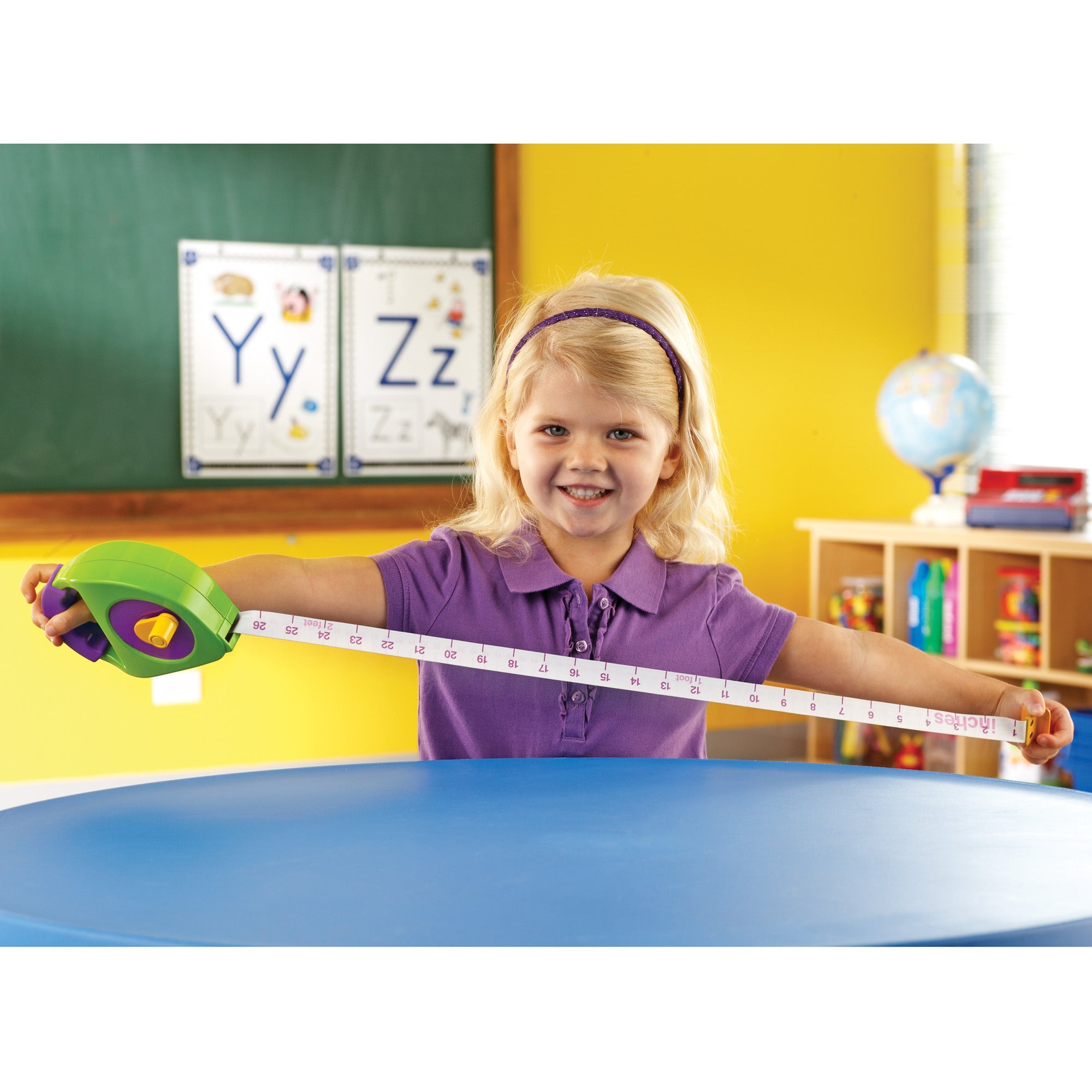 A child smiling while stretching out a chunky green and purple tape measure across a blue table in a classroom. The tape displays clear imperial and metric markings.