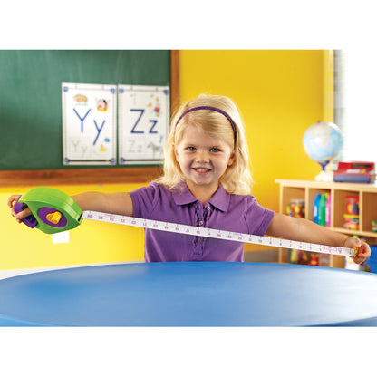 A child smiling while stretching out a chunky green and purple tape measure across a blue table in a classroom. The tape displays clear imperial and metric markings.