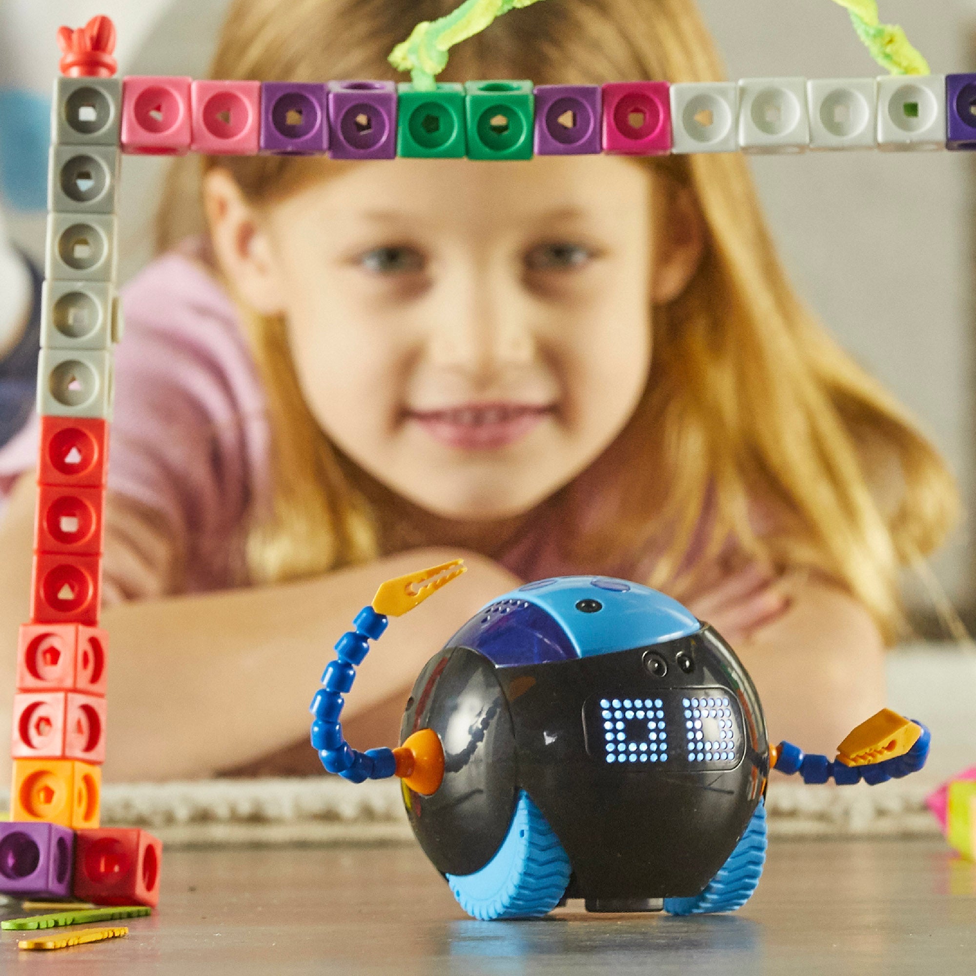 A child watches Cooper™ the STEM Robot navigate through a colourful archway made of building blocks. The spherical blue and black robot features glowing LED eyes and posable arms.