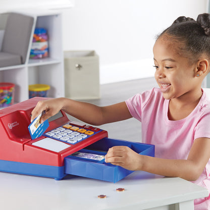 A child smiling while using a red and blue toy cash register. They are swiping a play credit card and opening the cash drawer, which contains realistic UK play money and coins.