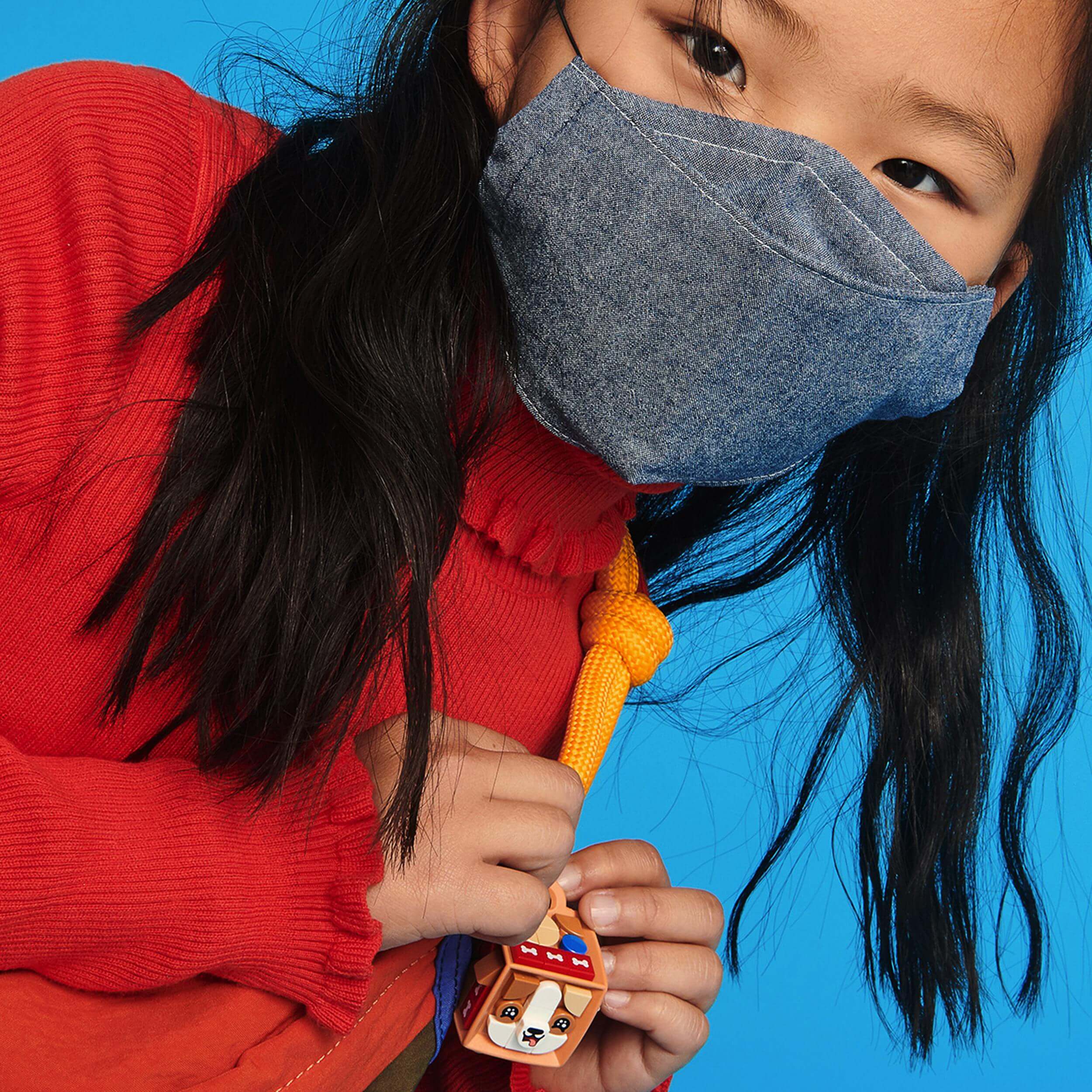 Child wearing a denim mask holding a colorful dog-themed bag tag, set against a bright blue background.