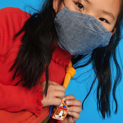 Child wearing a denim mask holding a colorful dog-themed bag tag, set against a bright blue background.