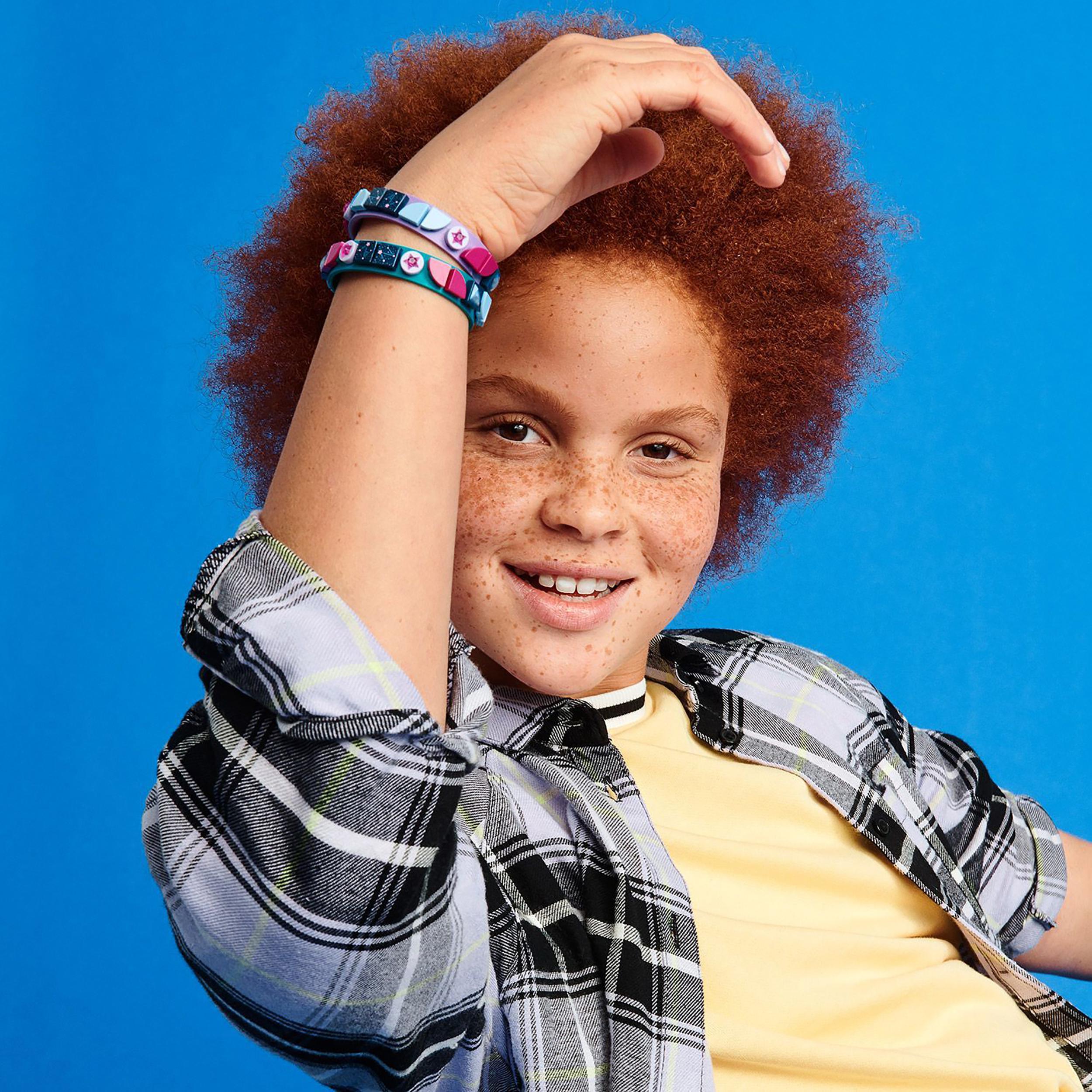 Child showcasing LEGO DOTS Starlight Bracelets with teal and purple tiles against a bright blue background.