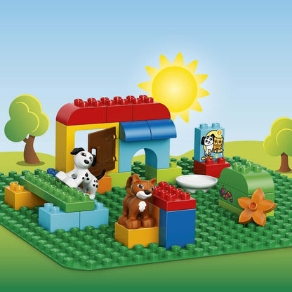 Colorful LEGO® DUPLO scene with a house, dog, cat, and various building elements on a green baseplate under a sunny sky.