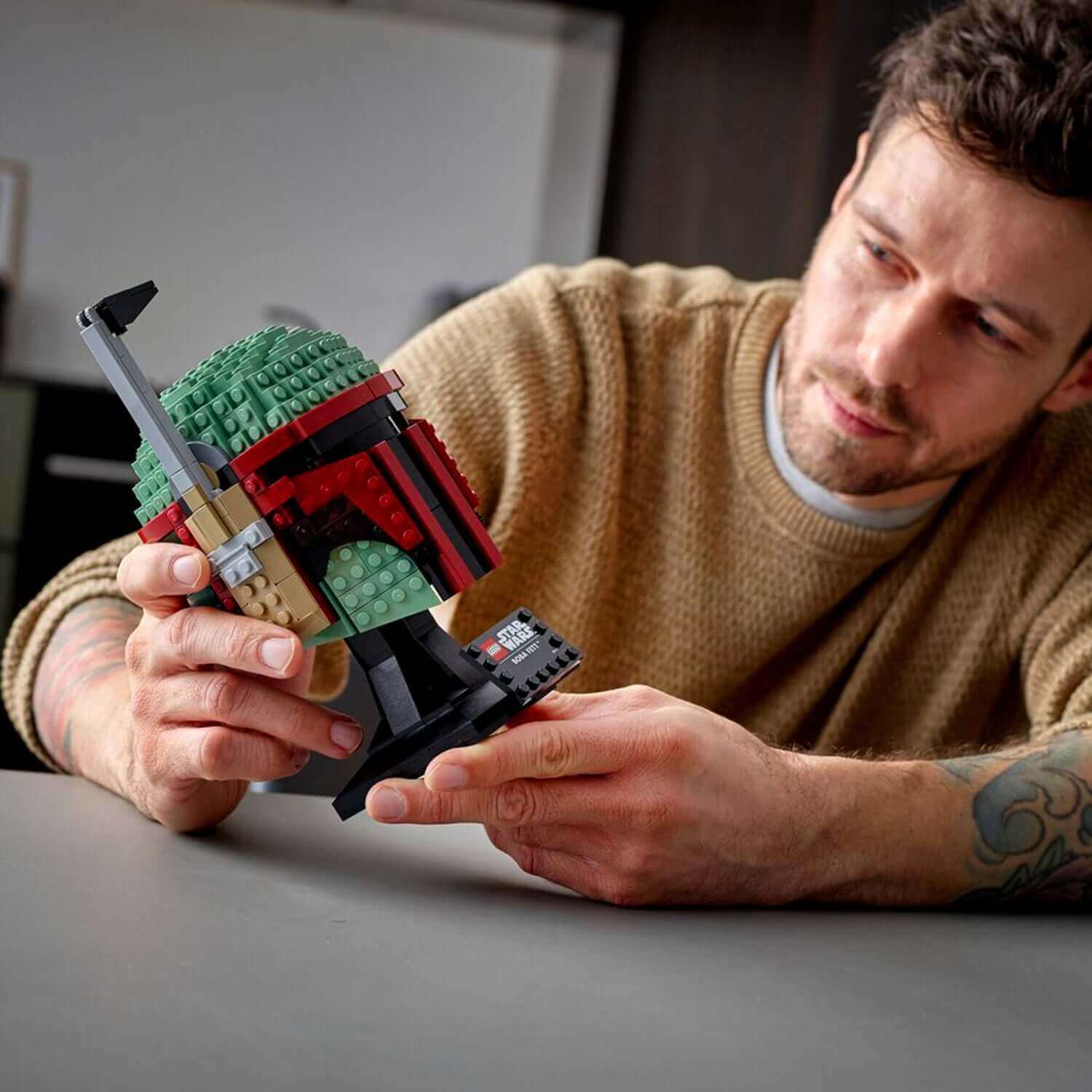 Adult builder showcasing the LEGO® Star Wars Boba Fett Helmet Building Set 75277 on a table.
