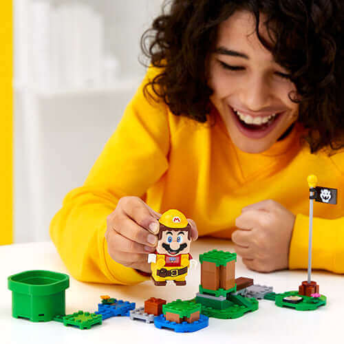 Child in a yellow sweater interacting with LEGO® Super Mario™ Builders set and Power-Up Pack, smiling with Mario figure.