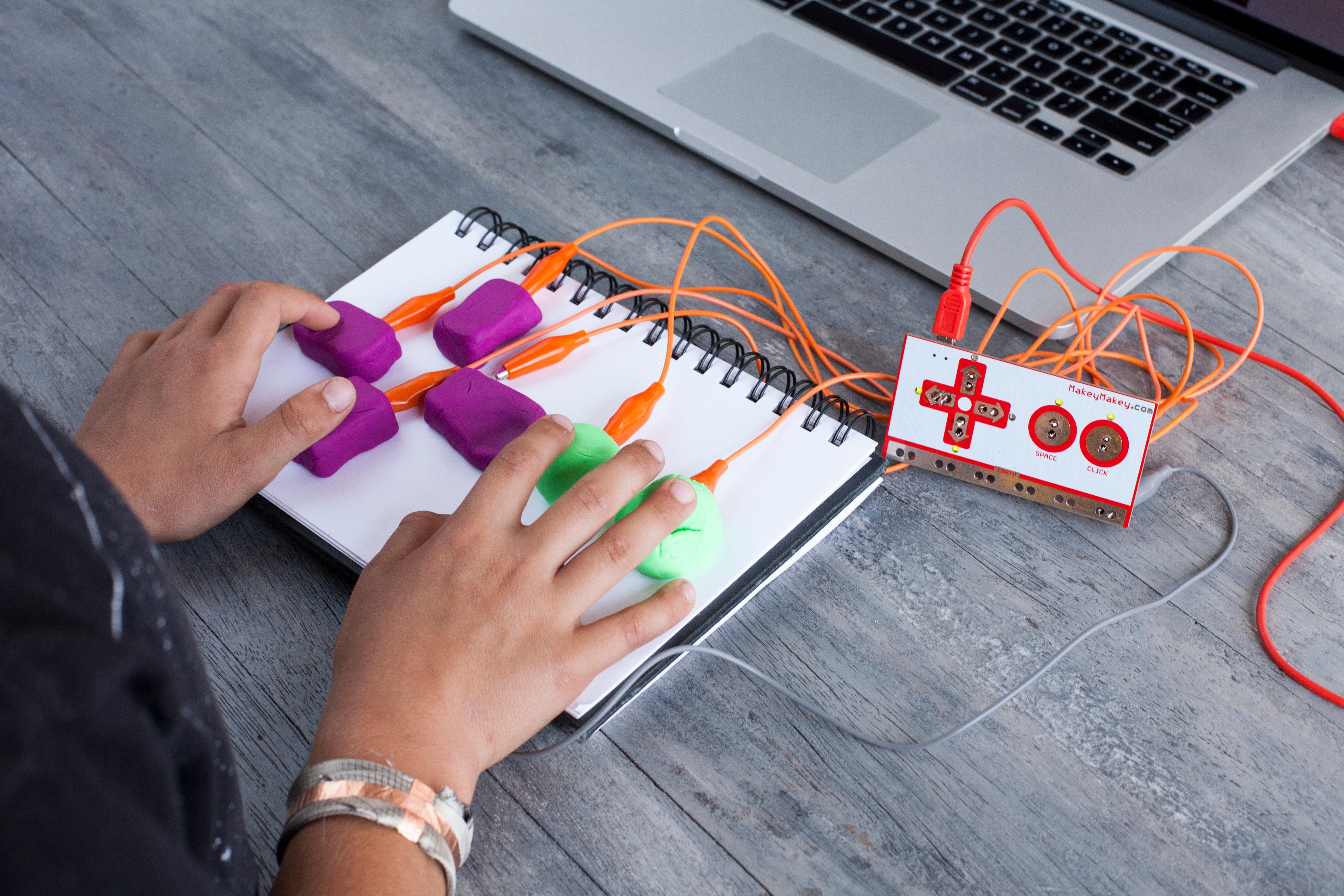 Hands using colorful buttons on a notepad, connected to a computer via wires, showcasing interactive design.