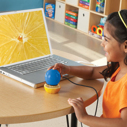 A young girl in an orange shirt smiles as she uses the blue Zoomy™ 2.0 Handheld Digital Microscope to examine the internal structure of a lemon half. The egg-shaped microscope is held directly against the fruit, and a laptop screen displays a crystal-clear, 54x magnified view of the lemon's juice vesicles. The image, set in a classroom environment, demonstrates the device's ease of use and its ability to provide a new perspective on everyday objects for children aged 4 and up.