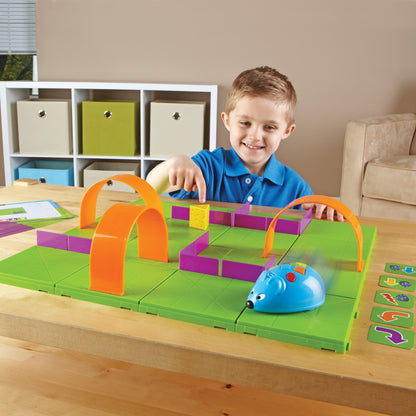 A child smiles while using the Code & Go® Robot Mouse Activity Set. "Colby," a blue robot mouse, navigates a custom green grid maze with orange tunnels and purple walls toward a plastic wedge of cheese.