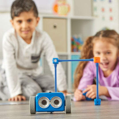 Two children playing with Botley 2.0 on a floor. The blue coding robot is in the foreground, having navigated through a blue and orange plastic obstacle.