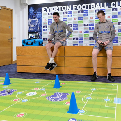 Two athletes seated on a bench with an Everton Football Club backdrop, interacting with a coding sports mat.