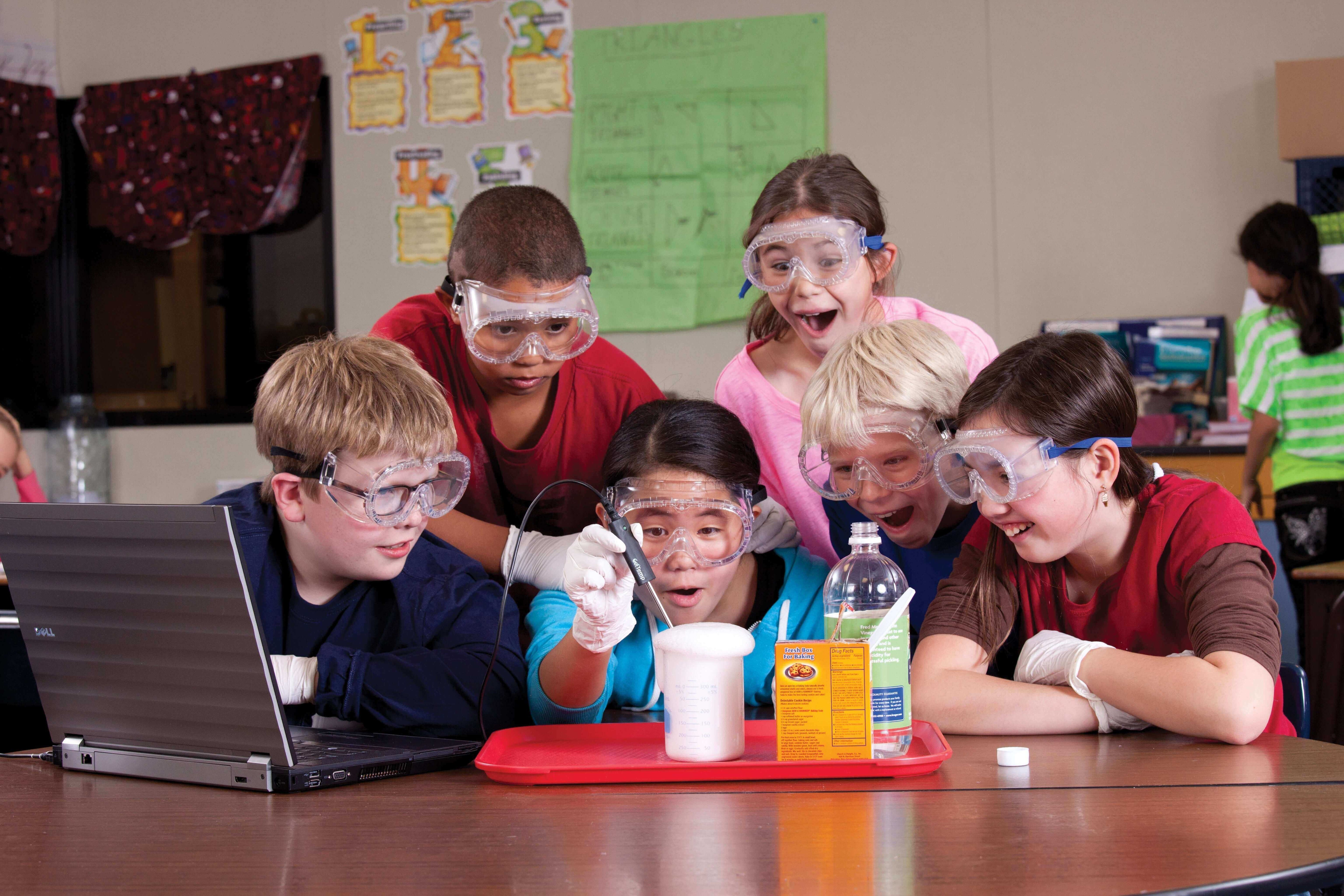 Students in safety goggles engaged in a science experiment, gathered around a laptop and a tray with materials.