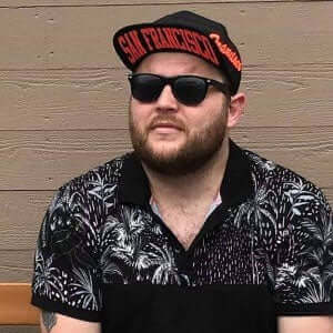 Man wearing sunglasses and a San Francisco hat, seated against a wooden backdrop, showcasing a stylish tropical shirt.
