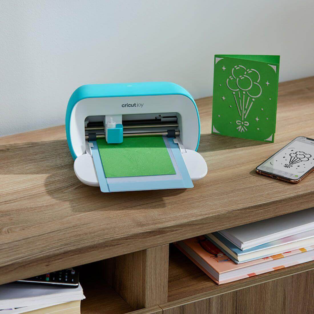 Cricut Joy cutting machine with green insert card in progress and a smartphone on a wooden desk.