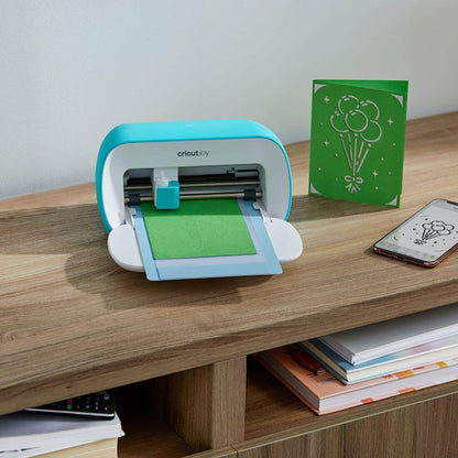 Cricut Joy cutting machine with green insert card in progress and a smartphone on a wooden desk.