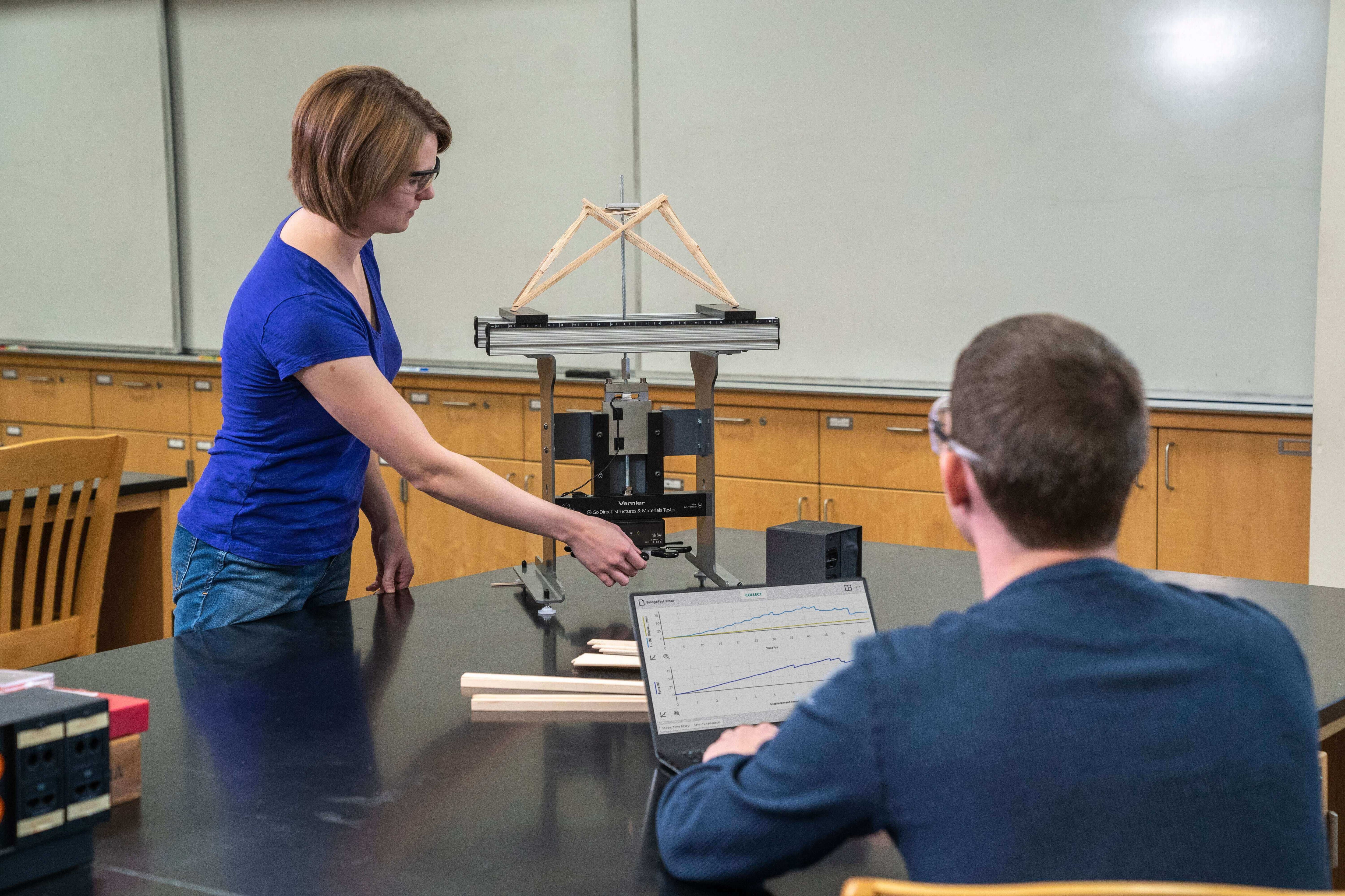 Student demonstrates a physics experiment with a bridge model in a classroom, while another student observes on a laptop.