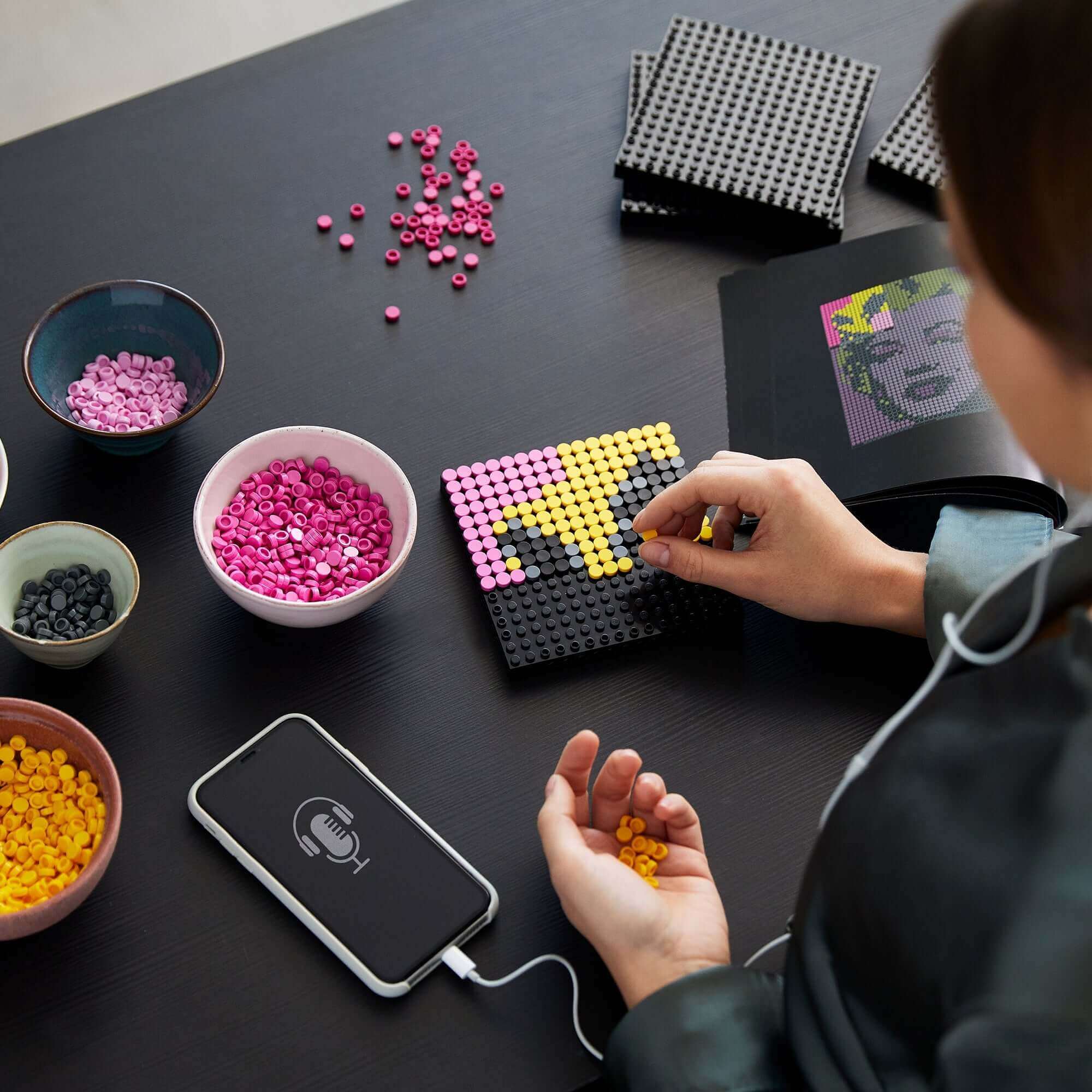 Person creating LEGO® Art of Andy Warhol's Marilyn Monroe with colorful bricks and smartphone on a table.