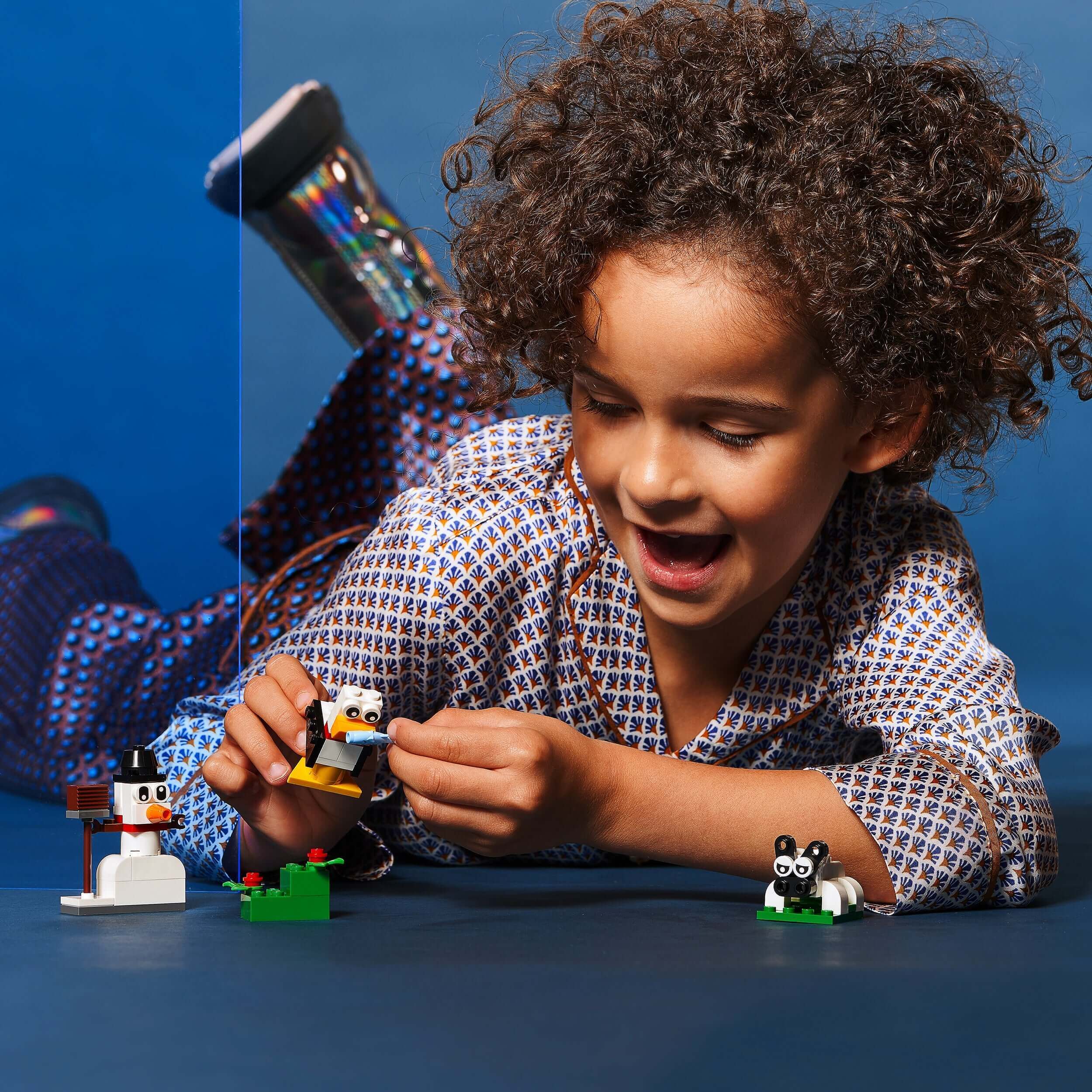 Child playing with LEGO® Classic Creative White Bricks Set 11012, building a snowman and animal figures on a blue background.