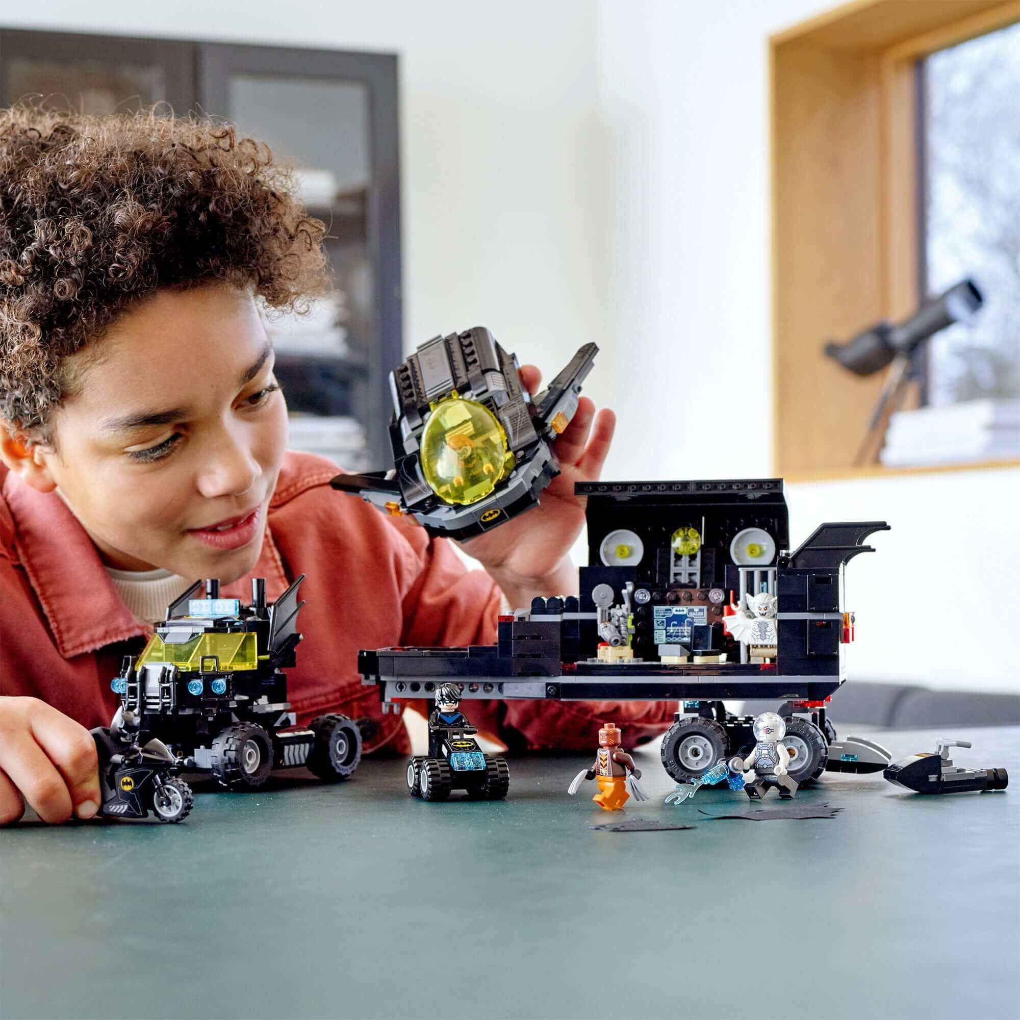 Child playing with LEGO® DC Batman Mobile Bat Base Truck Toy 76160, featuring vehicles and minifigures on a table.